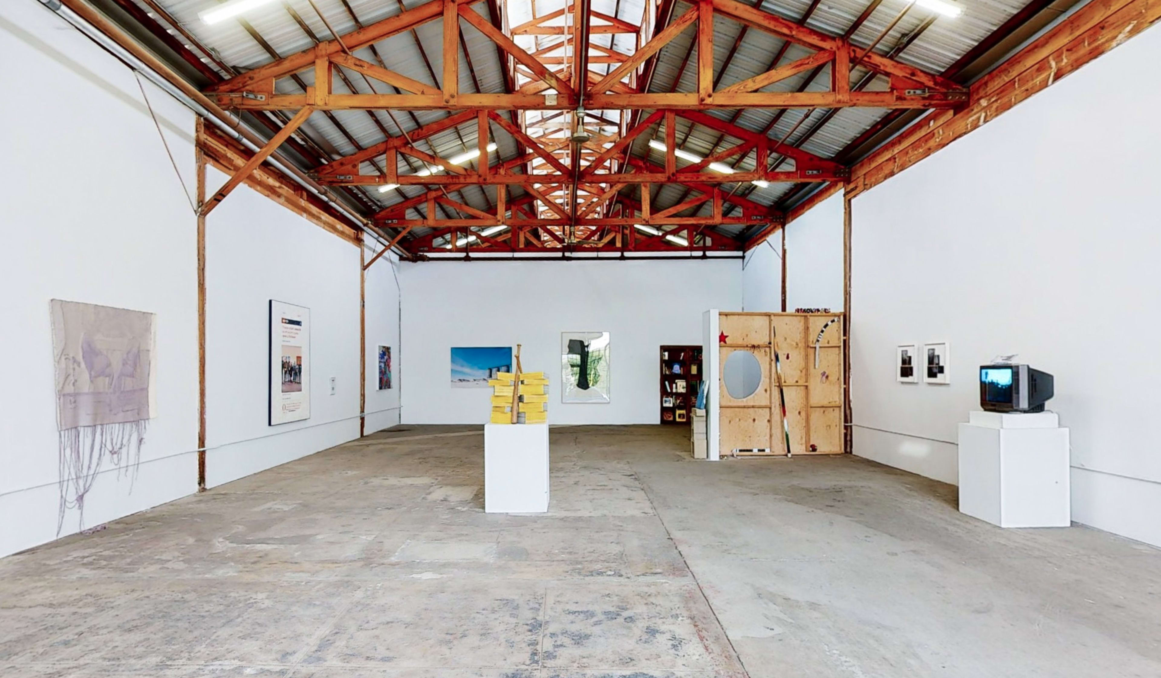 An art gallery with a high ceiling, featuring various artworks displayed on white walls and a wooden structure in the background.