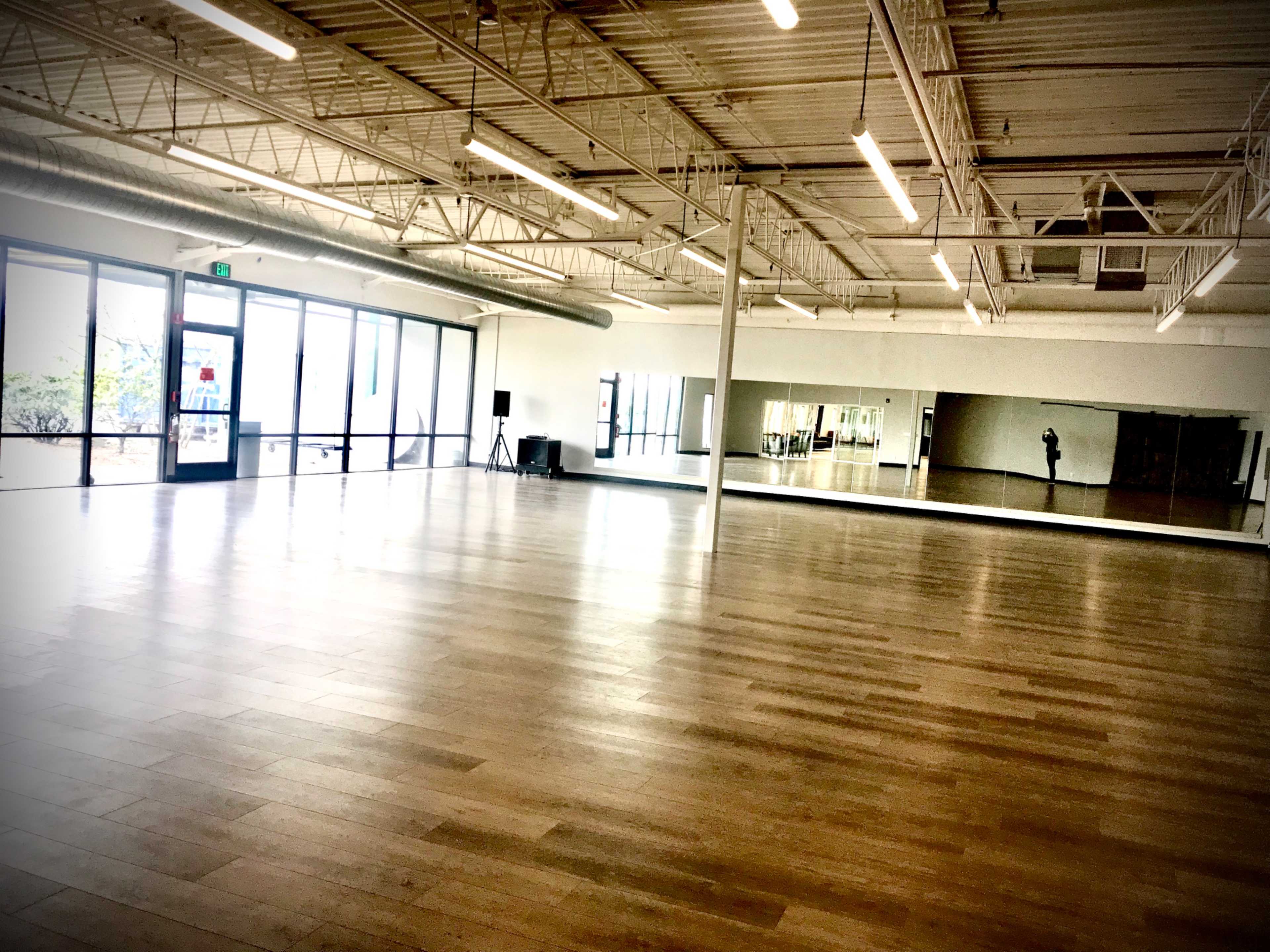 A large, empty dance studio with wooden floors, mirrors on one wall, and glass doors leading outside.