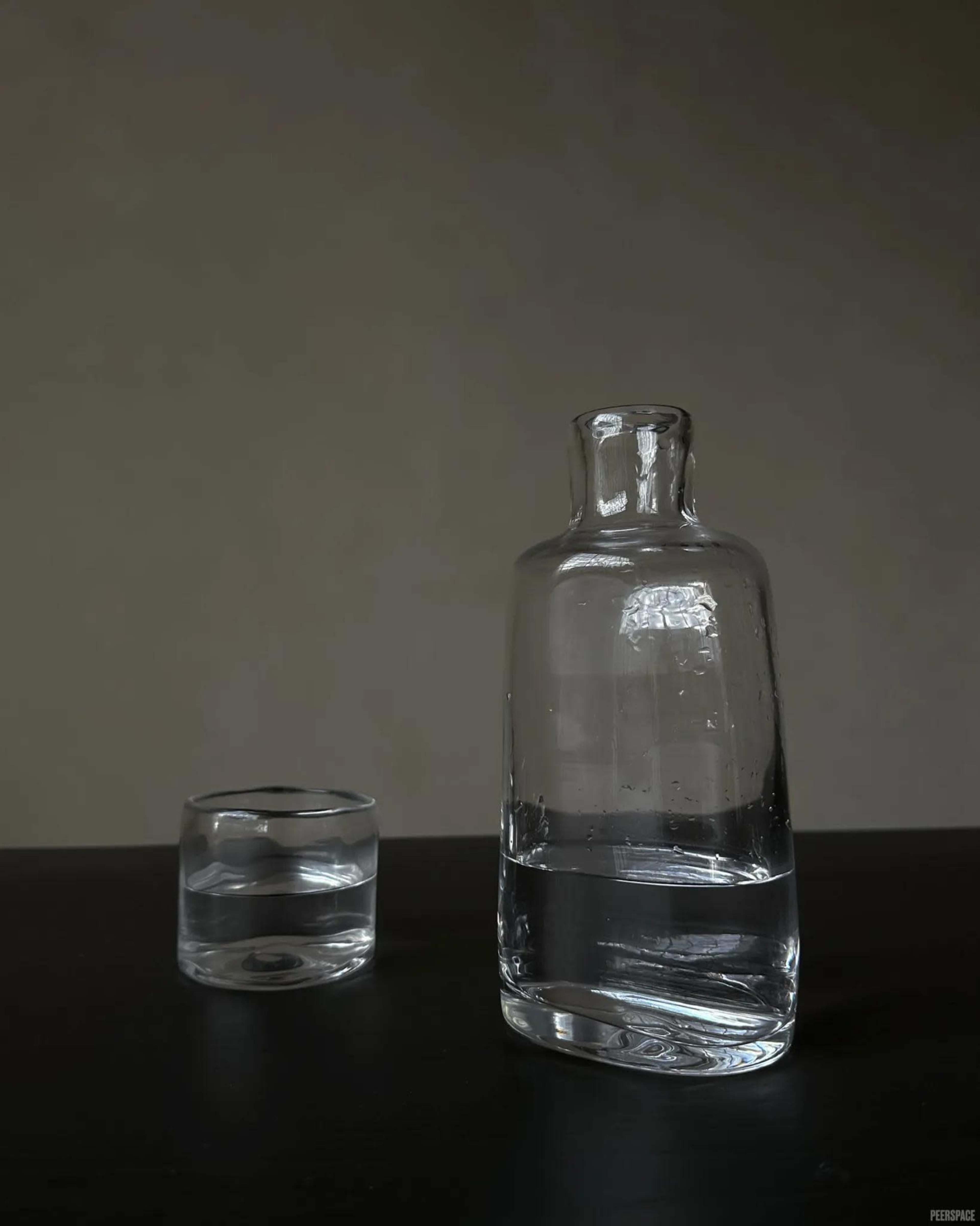 A clear glass bottle and a small glass containing water are placed on a dark surface against a neutral background.
