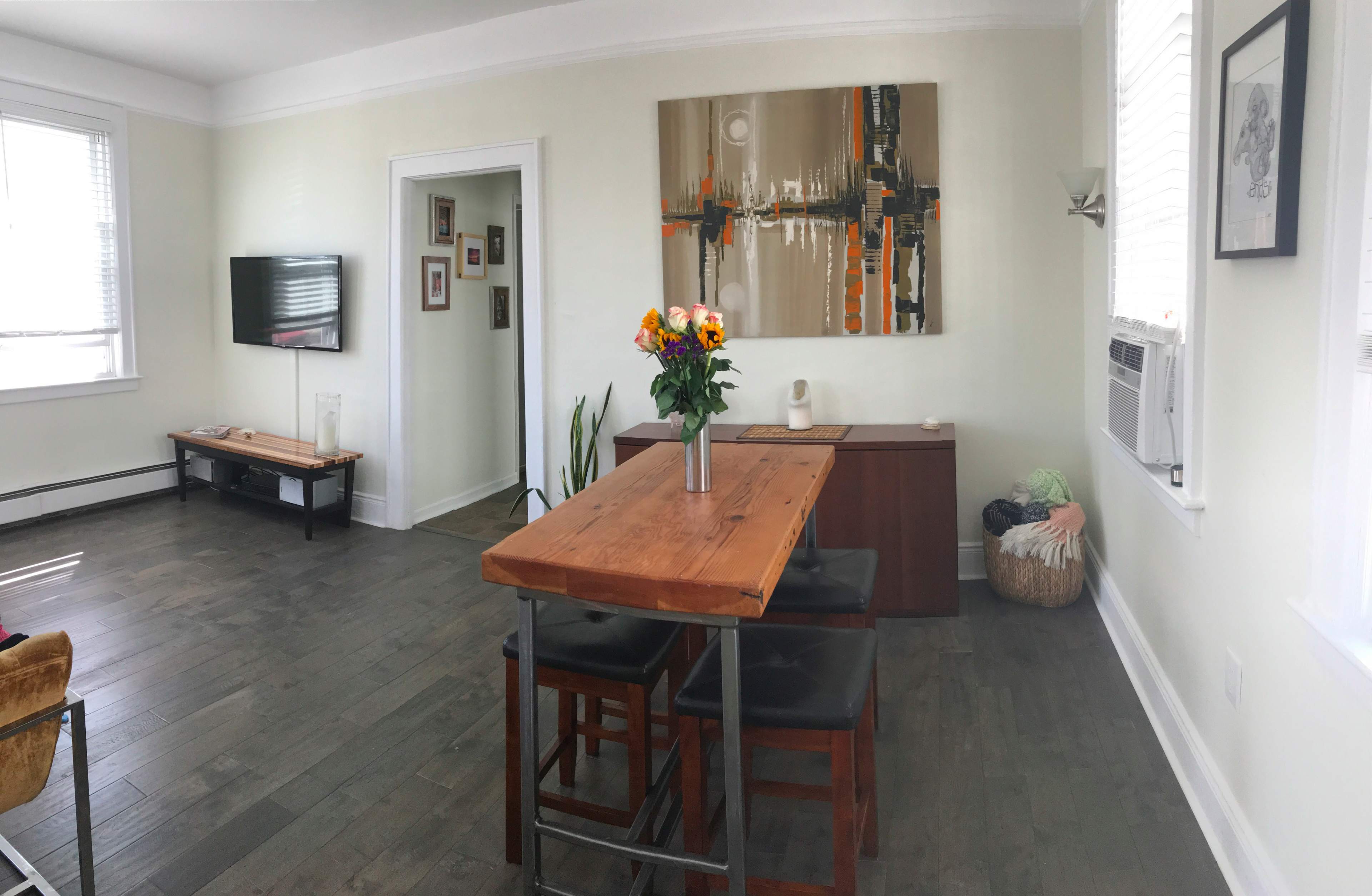 A dining area features a wooden table with black chairs, surrounded by minimalist decor, a painting on the wall, and a bouquet of flowers in a vase.