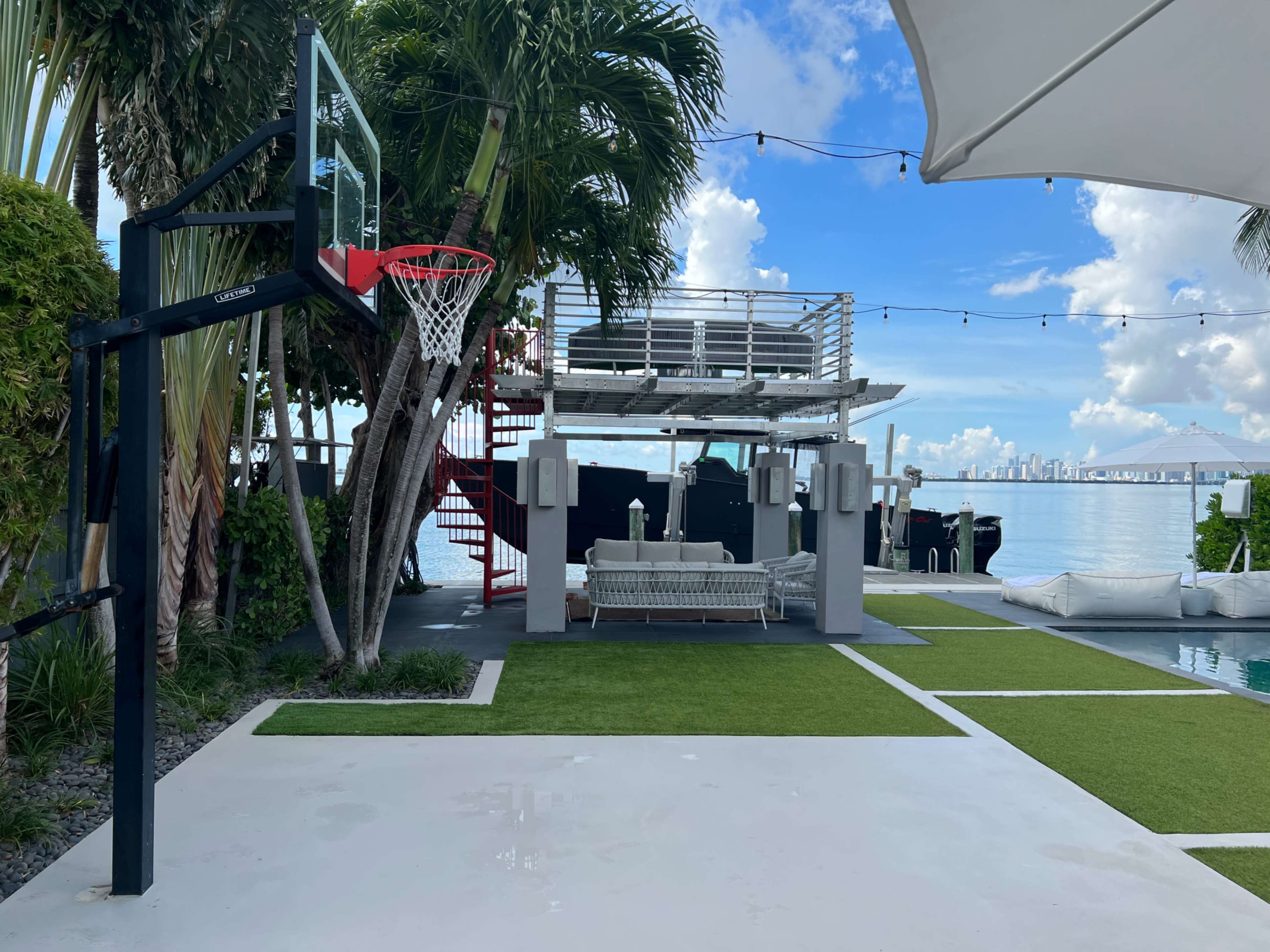 A modern outdoor space with a basketball hoop, a lounge area, and a view of a body of water and city skyline in the background.