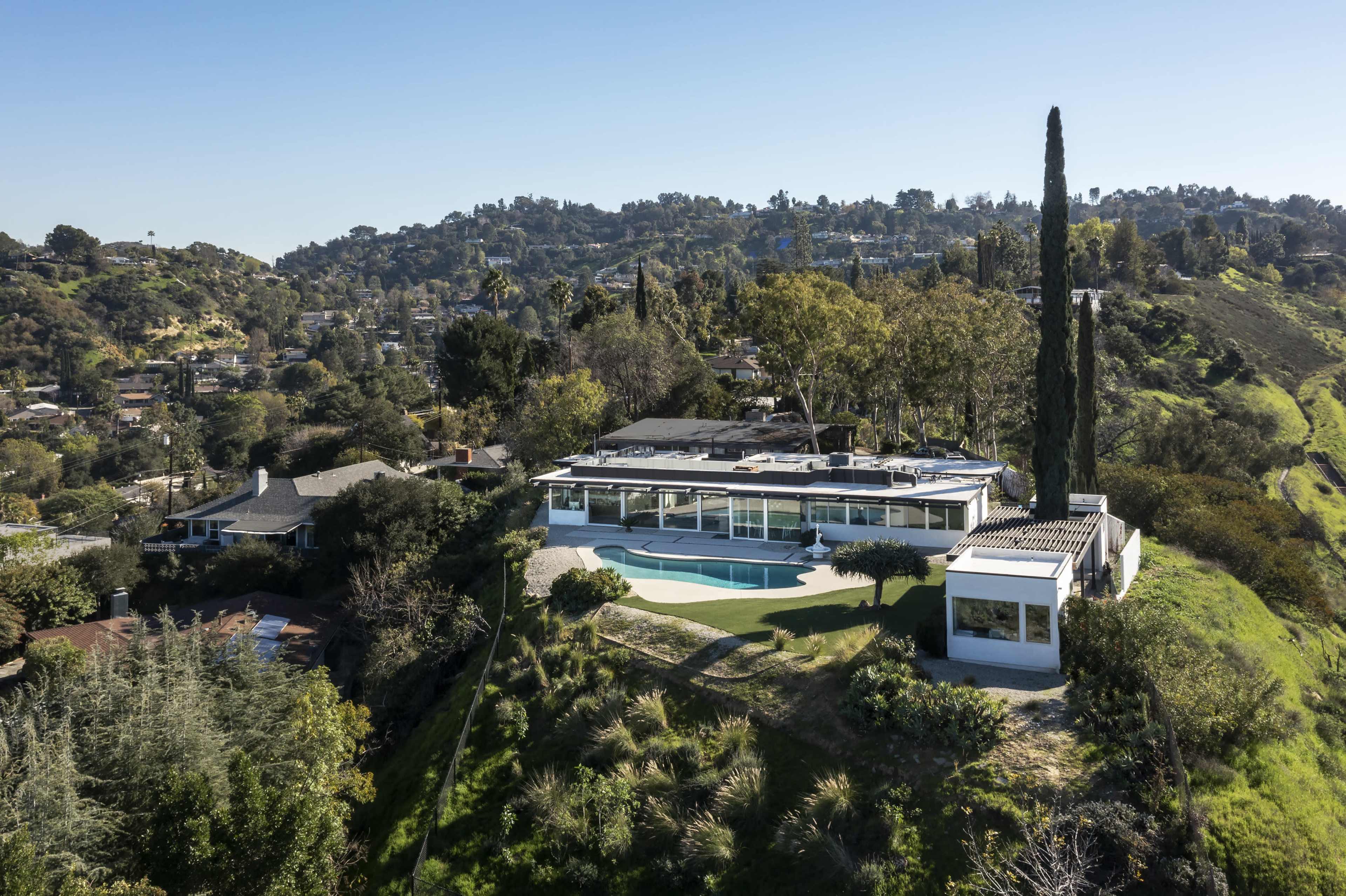The image shows a modern, single-story house with a pool, situated on a hillside surrounded by greenery and other residential buildings.