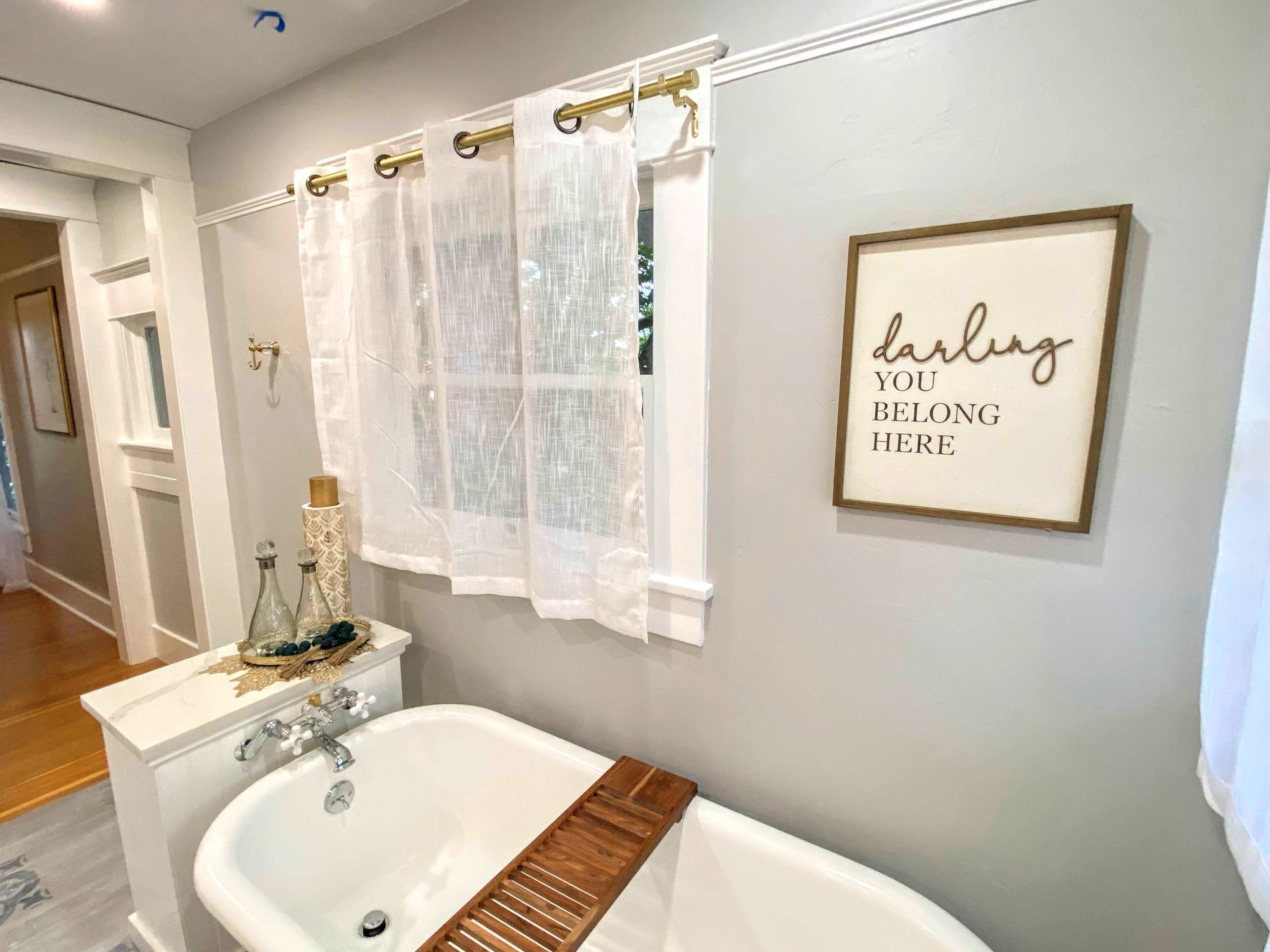 A bathroom with a vintage bathtub, light curtains over a window, and a framed sign on the wall that reads "darling you belong here."