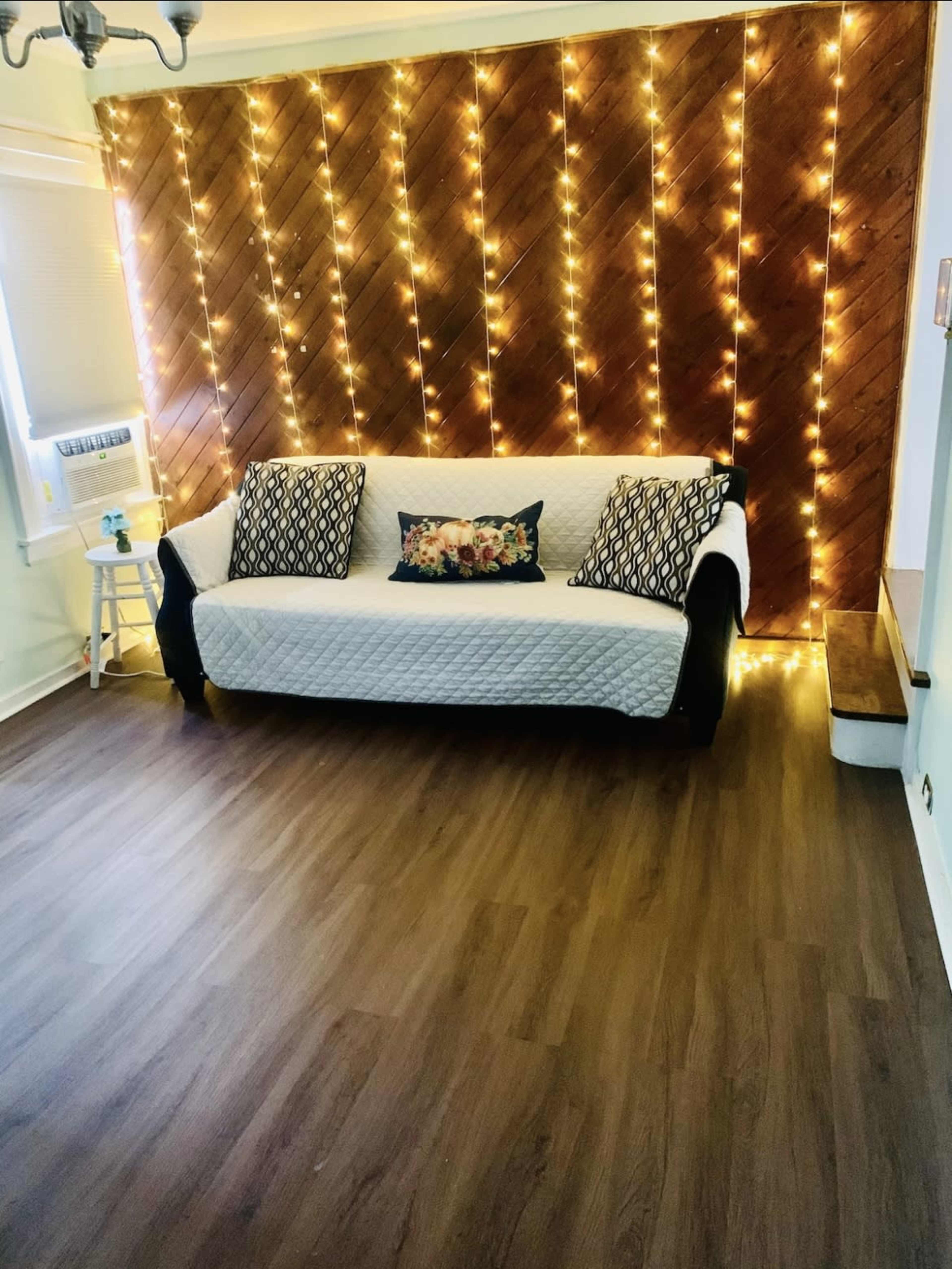 A white couch with patterned pillows sits against a wooden wall adorned with fairy lights in a brightly lit room with vinyl flooring.