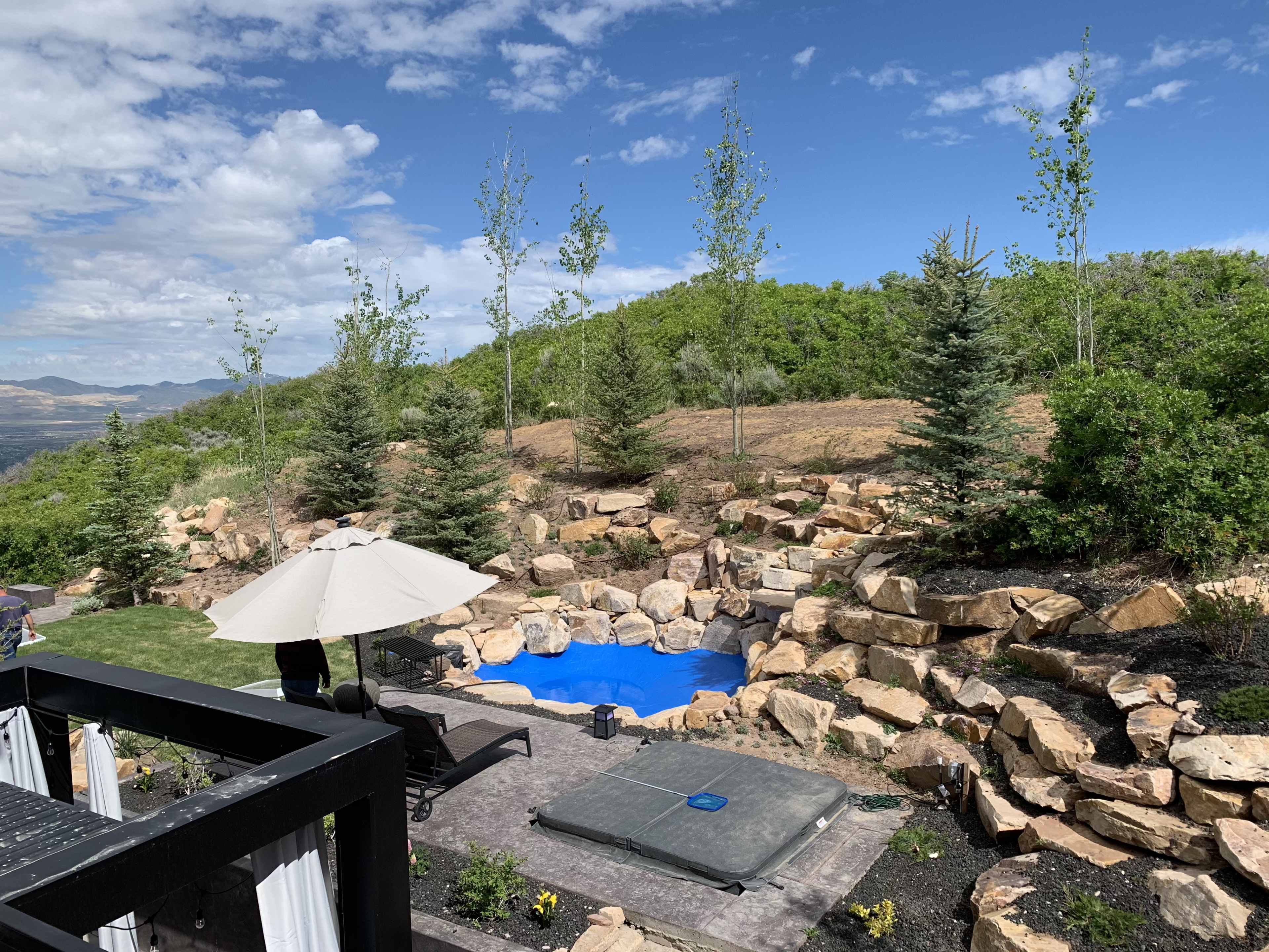 A landscaped backyard featuring a blue pool surrounded by large rocks, trees, and an umbrella over a lounge area.