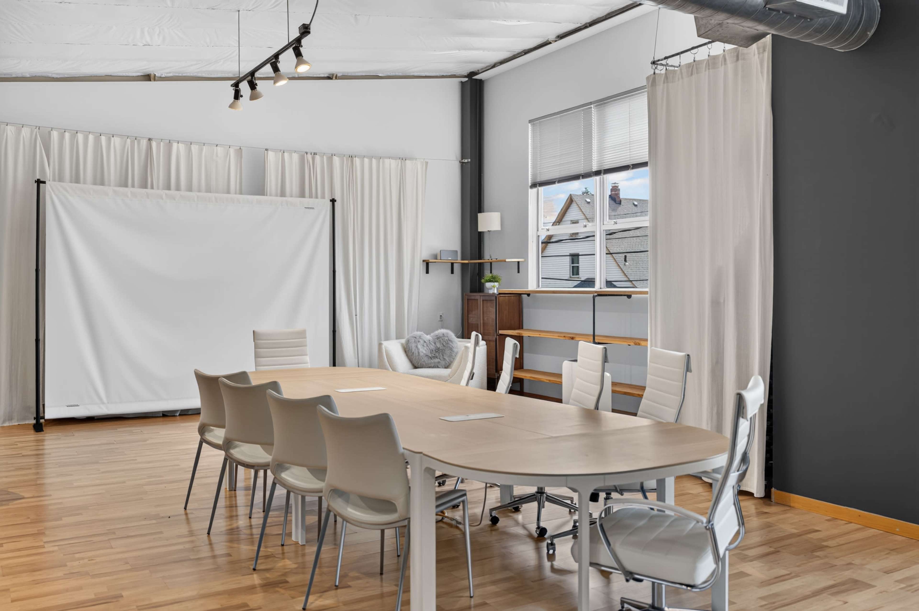 A spacious meeting room features a long wooden table surrounded by white chairs, with a projector screen and large windows in the background.