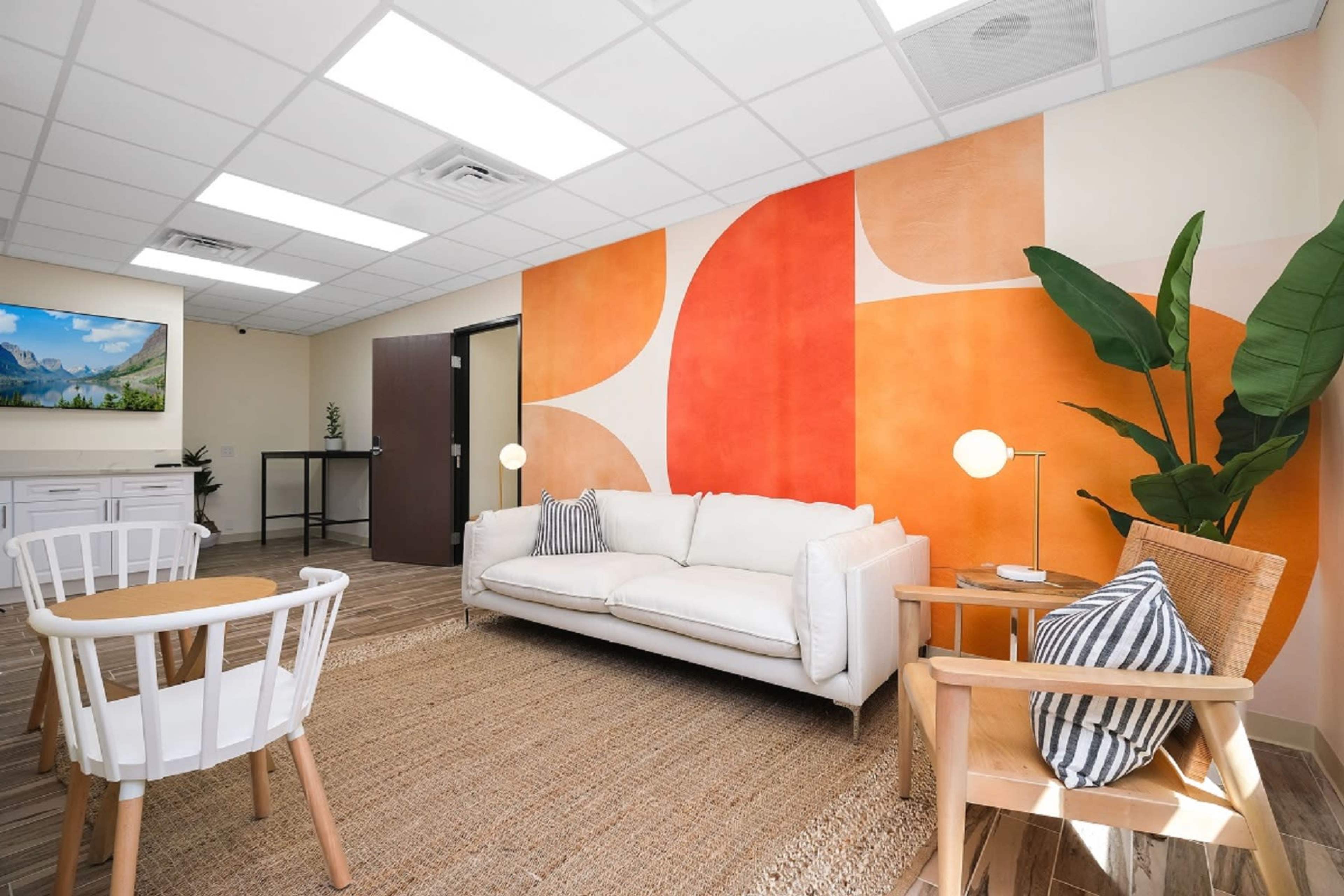 A modern waiting area with a white couch, two wooden chairs, a round rug, and a colorful accent wall featuring orange and cream shapes.
