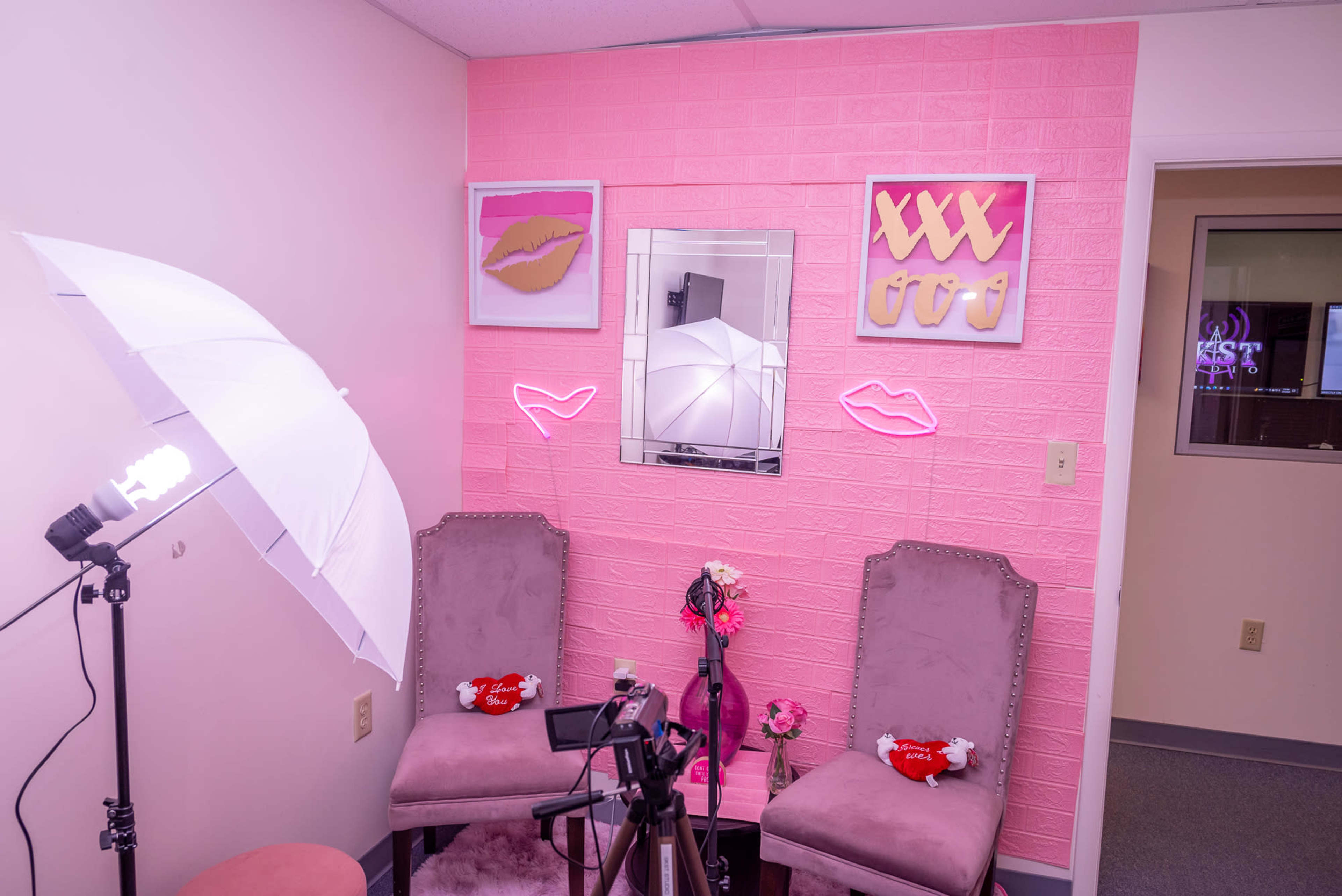 A pink room features two chairs, a mirror, and neon decorations, accompanied by photography equipment and soft lighting.