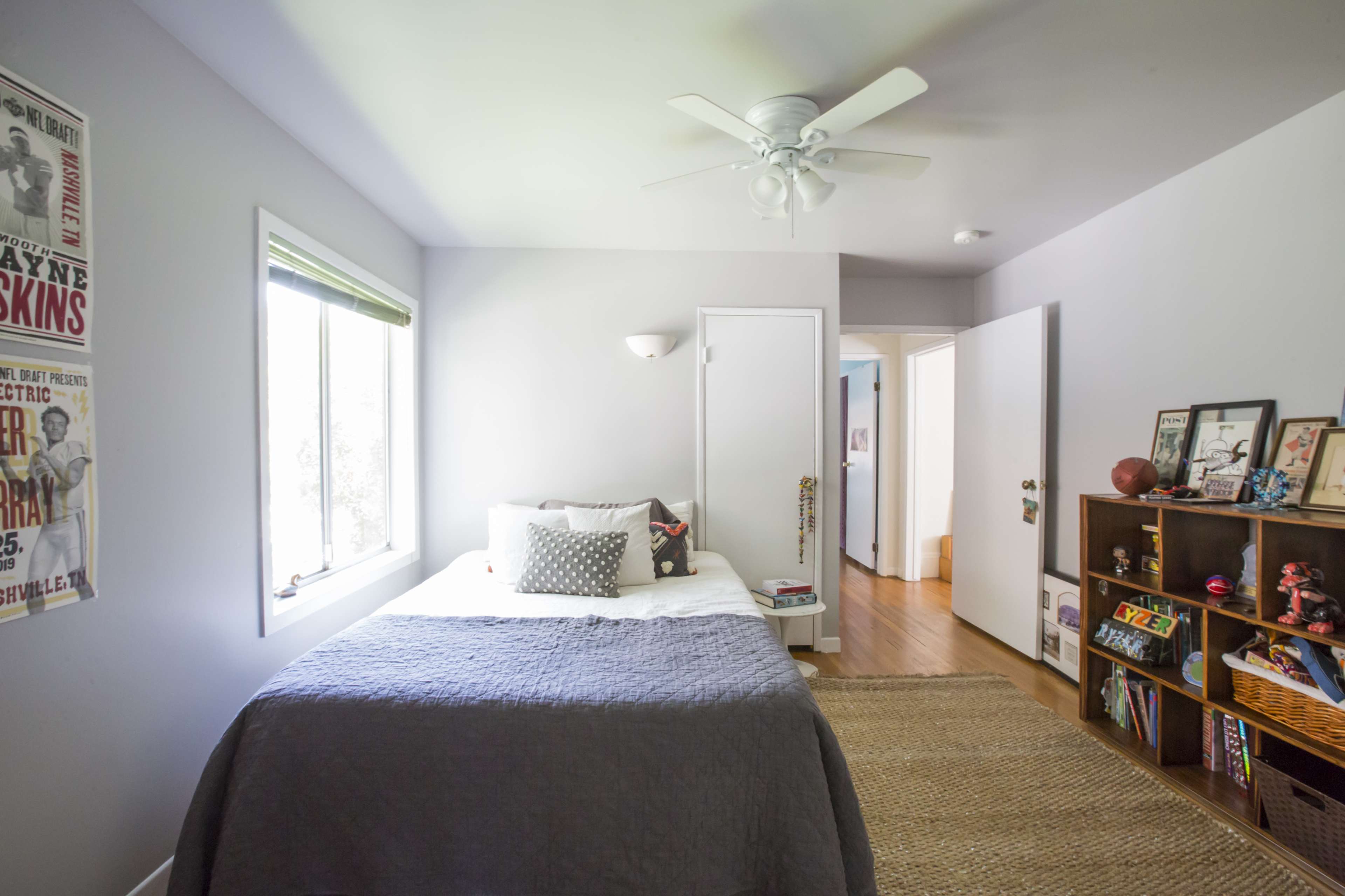 The image shows a simple bedroom featuring a bed with gray bedding, a window with blinds, a ceiling fan, and a wooden bookshelf filled with books and decorative items.