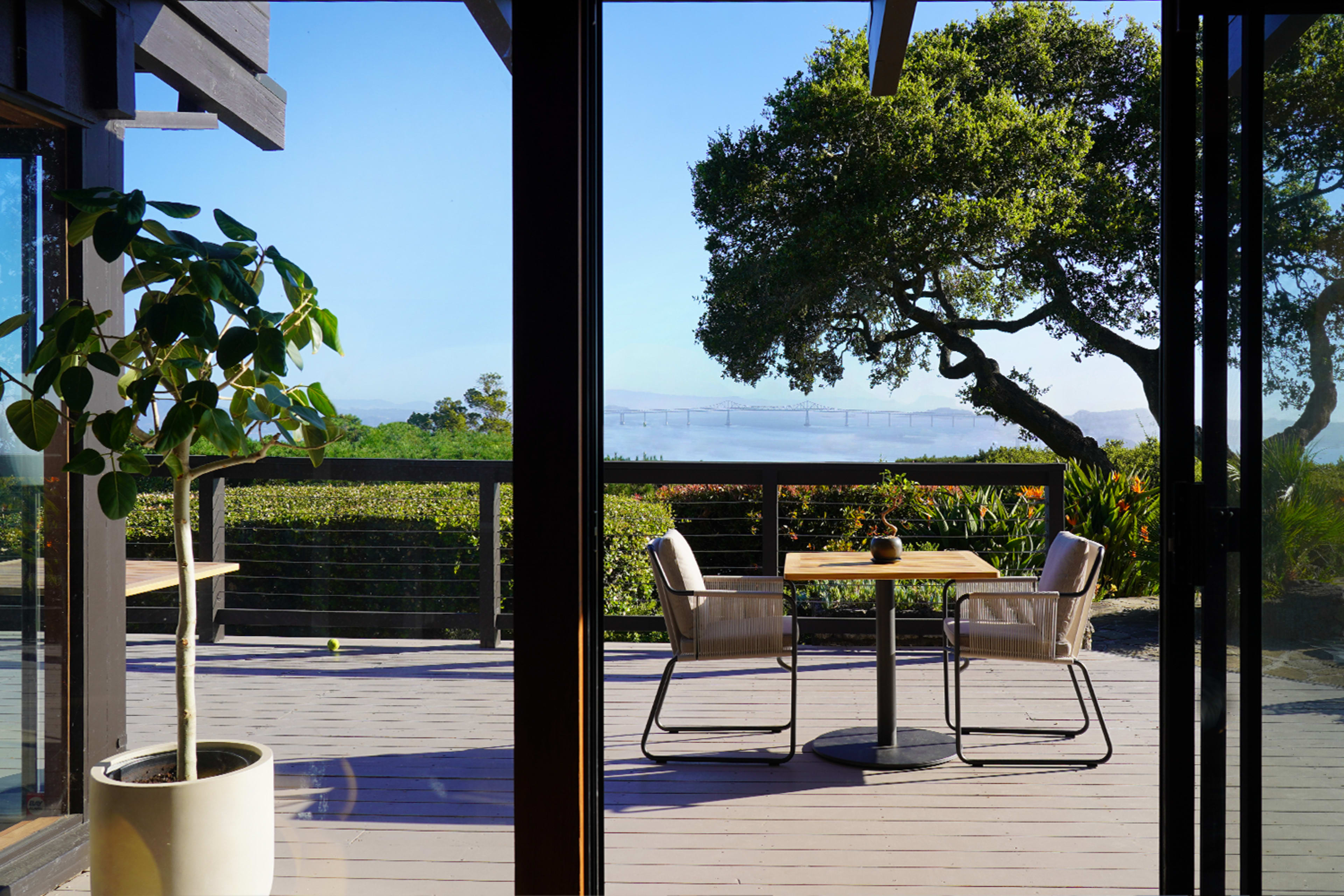 A table with two chairs is set on a wooden deck overlooking a landscape featuring greenery and a distant bridge.
