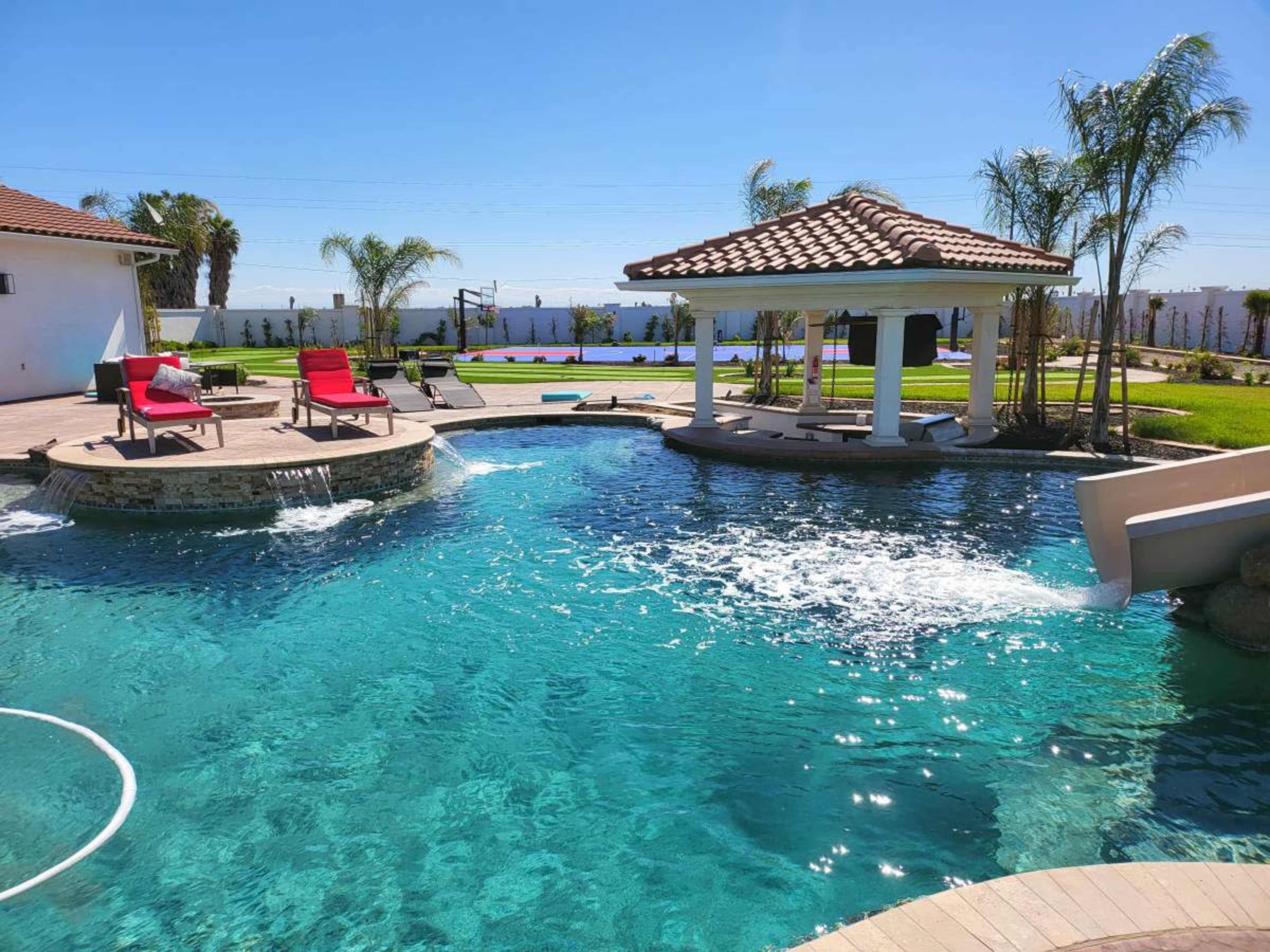 The image shows a landscaped pool area with a gazebo, lounge chairs, and palm trees under a clear blue sky.
