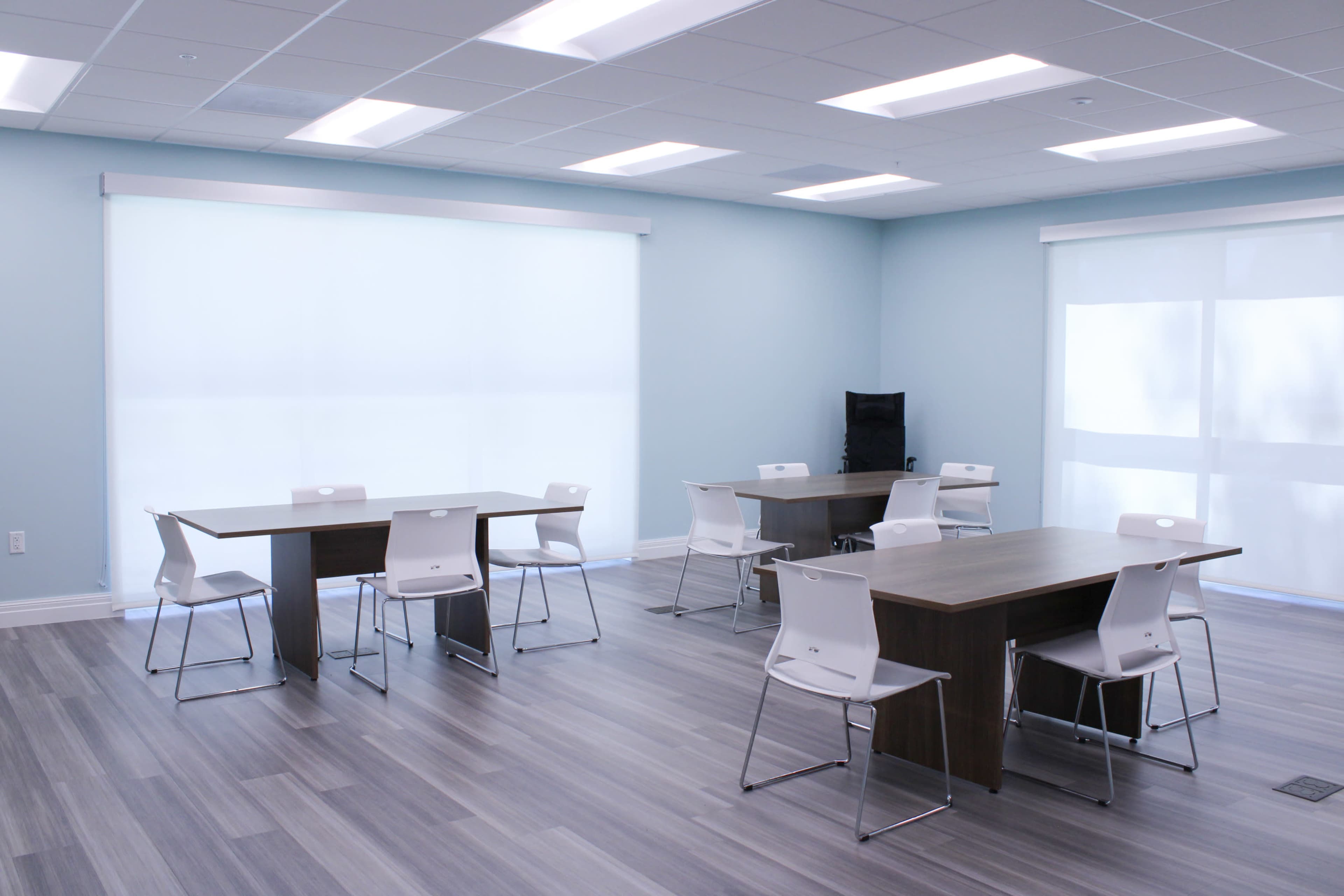 A modern classroom with several tables and chairs arranged on a wooden floor, illuminated by natural light from large windows.