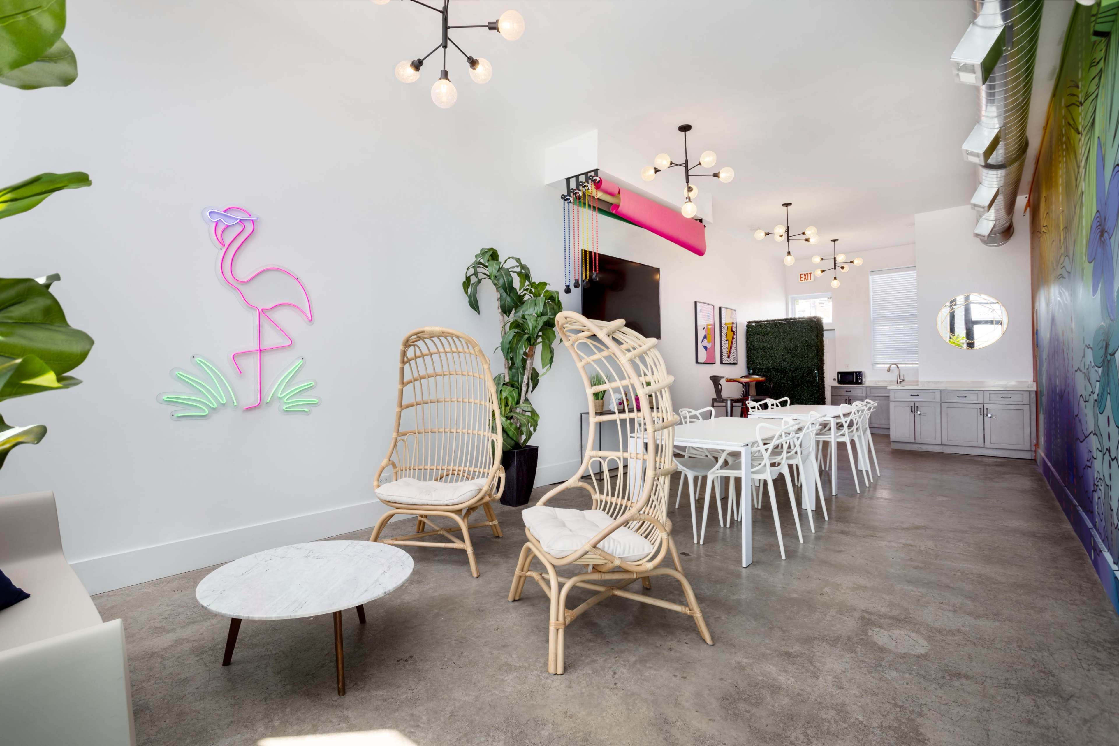 A modern interior space featuring rattan chairs, a neon flamingo wall decoration, and a long table with white chairs against a bright wall.