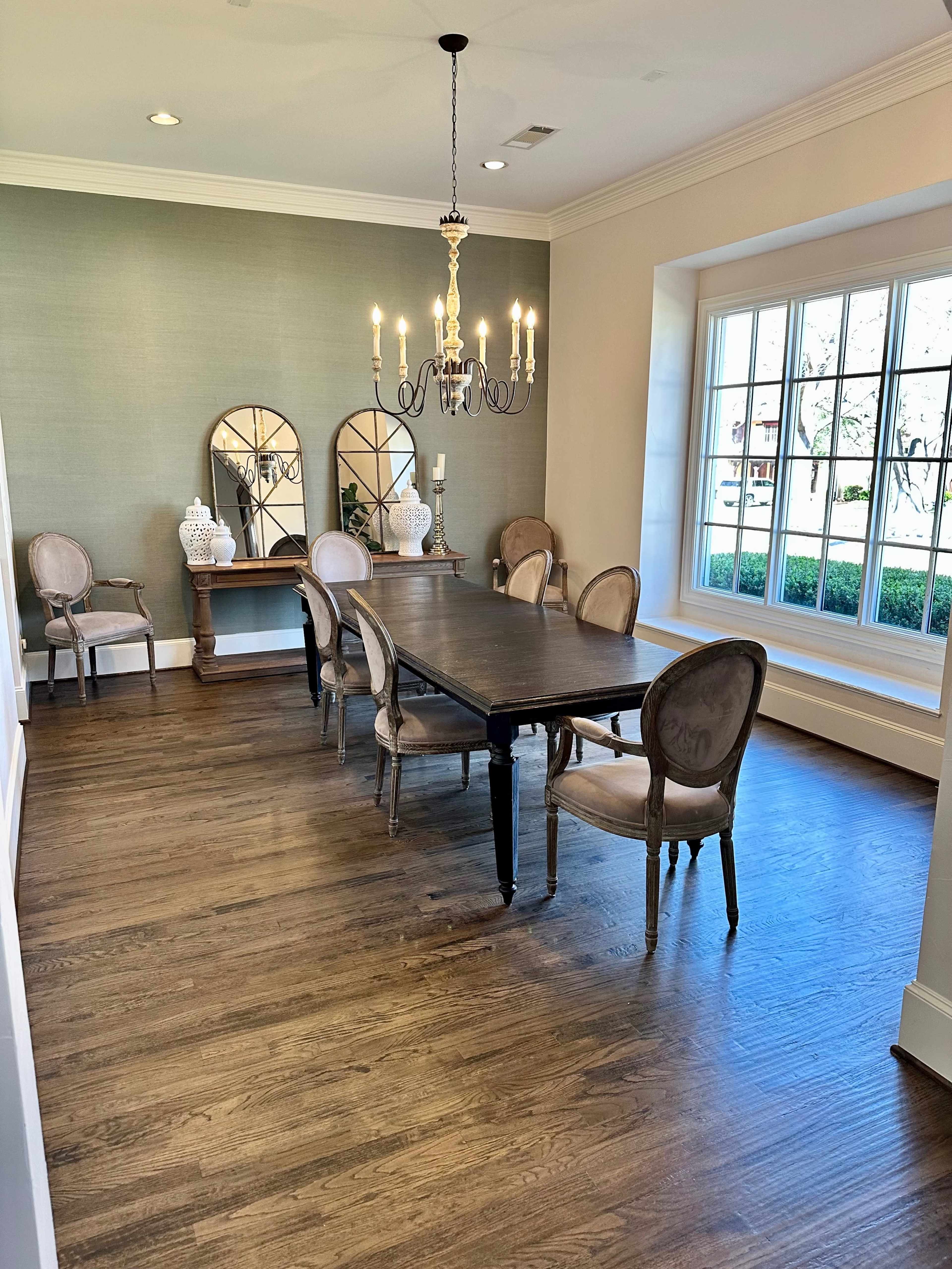 A dining room features a wooden table surrounded by six upholstered chairs, with a chandelier overhead and a side console with decorative mirrors and vases against the wall.