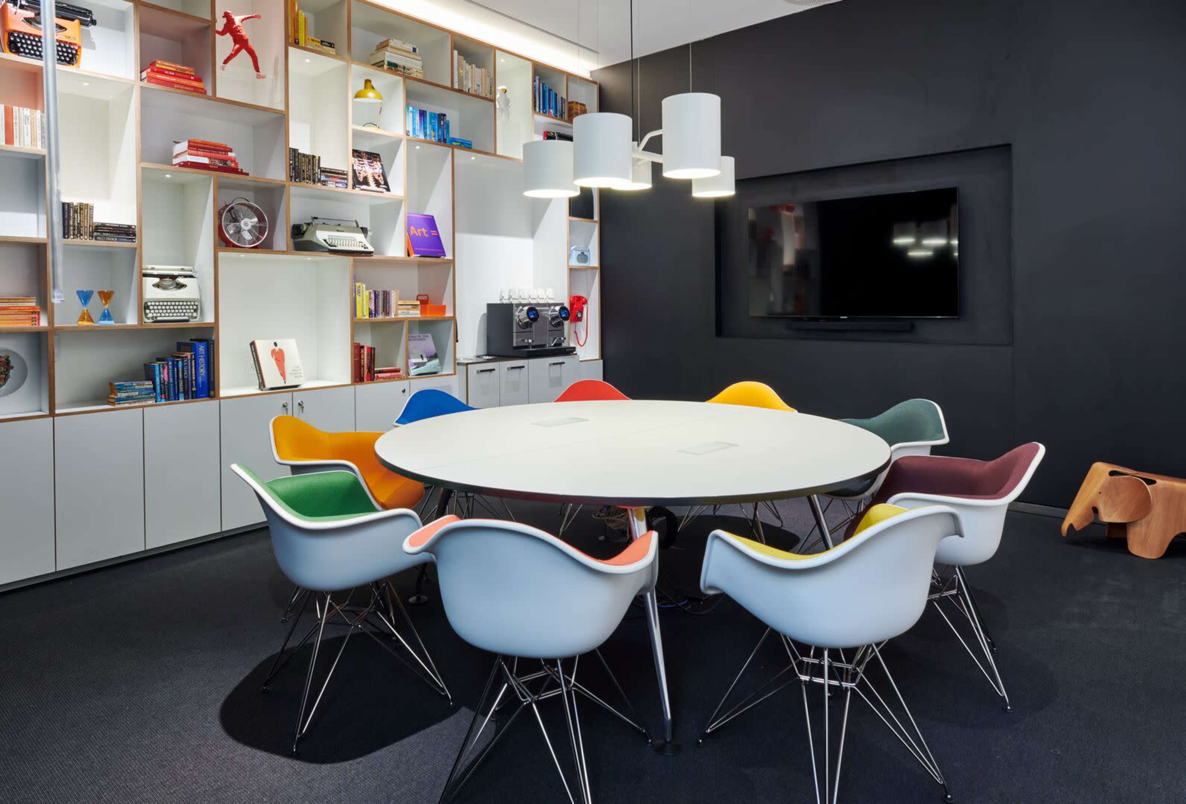 A modern conference room features a round table surrounded by colorful chairs, with shelves of books and decorative items in the background.