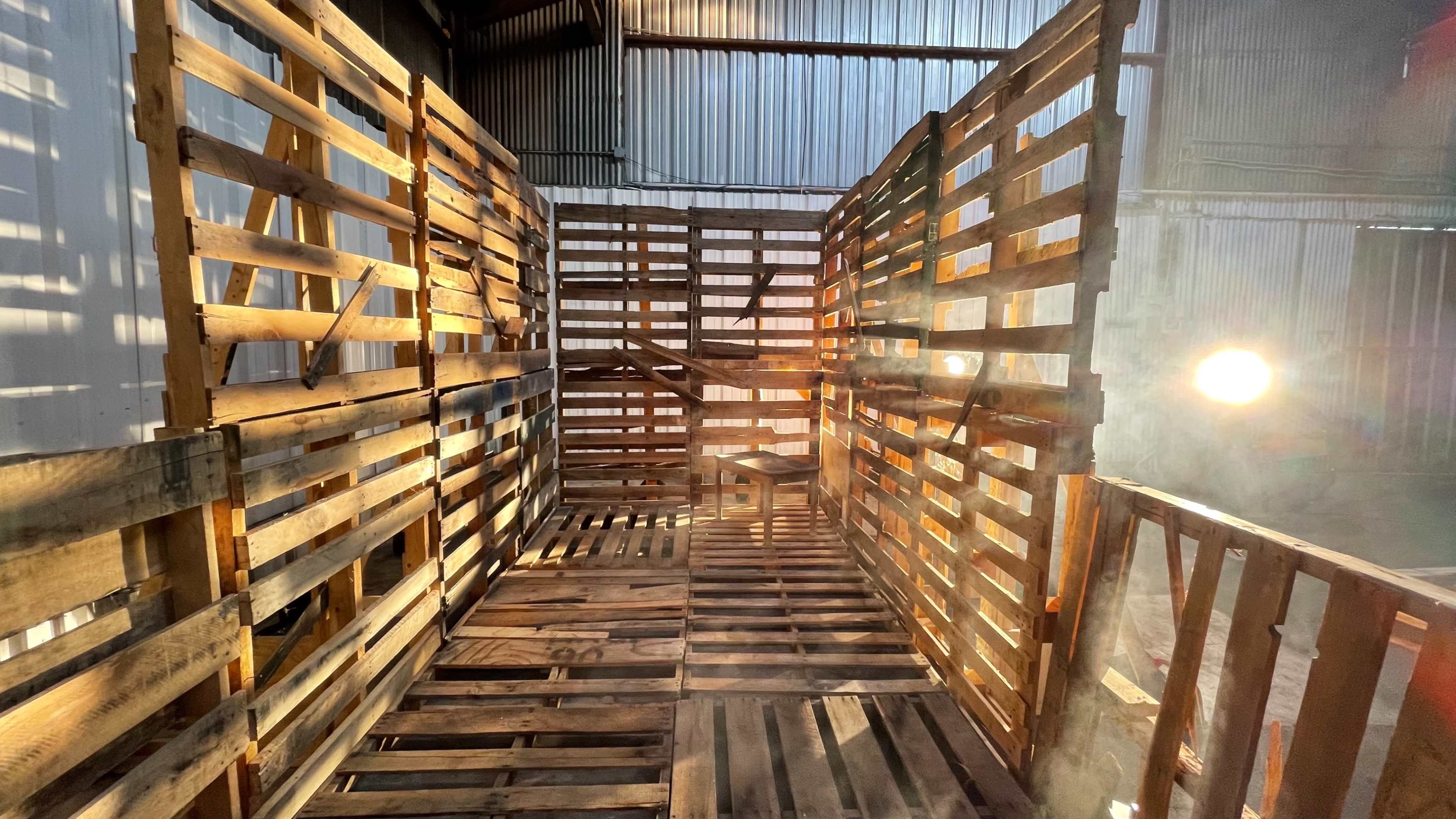 An interior space constructed from wooden pallets, featuring a walkway with a beam of light illuminating the area.