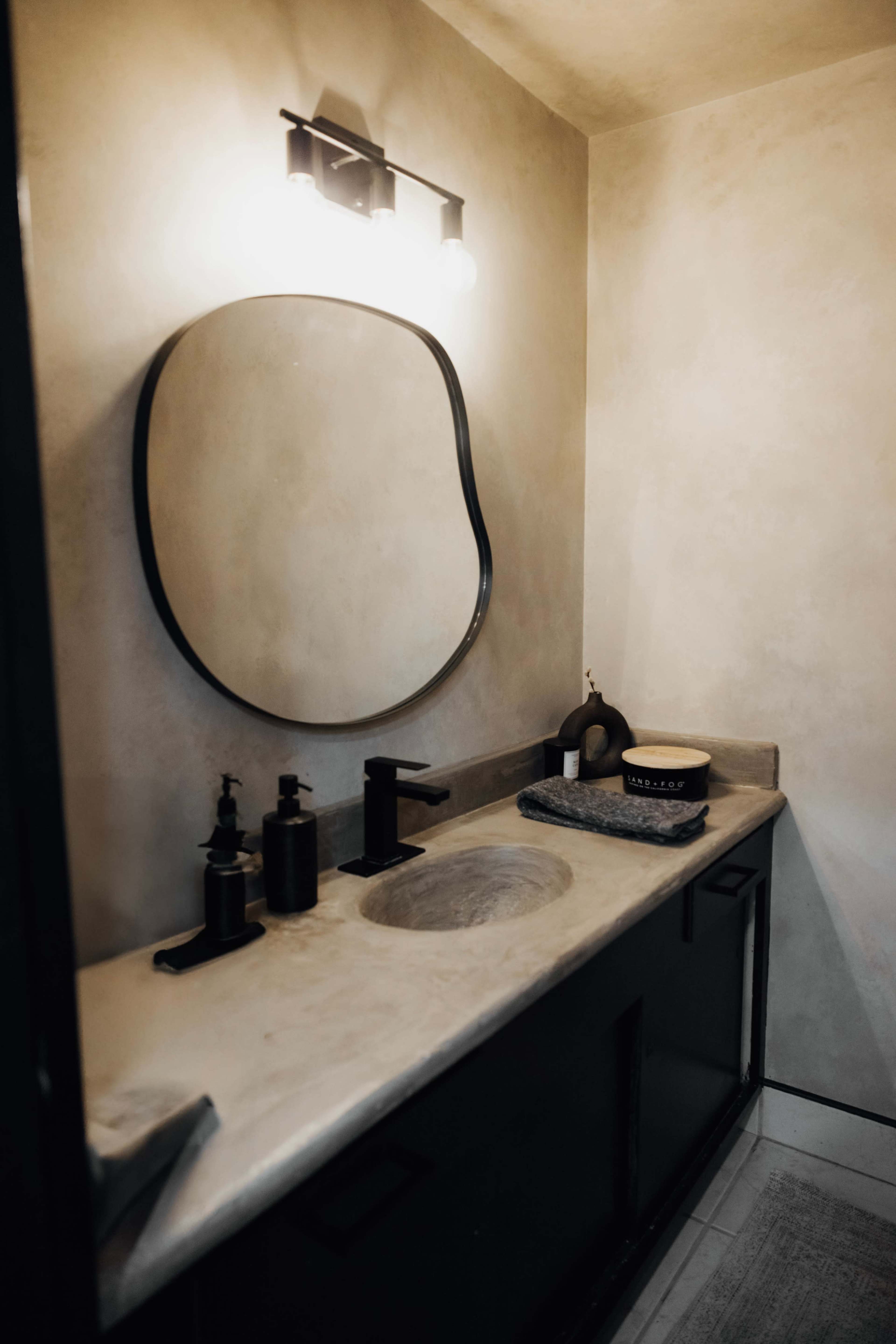 The image shows a modern bathroom with a round mirror, a sleek sink, and various personal care items on a countertop.