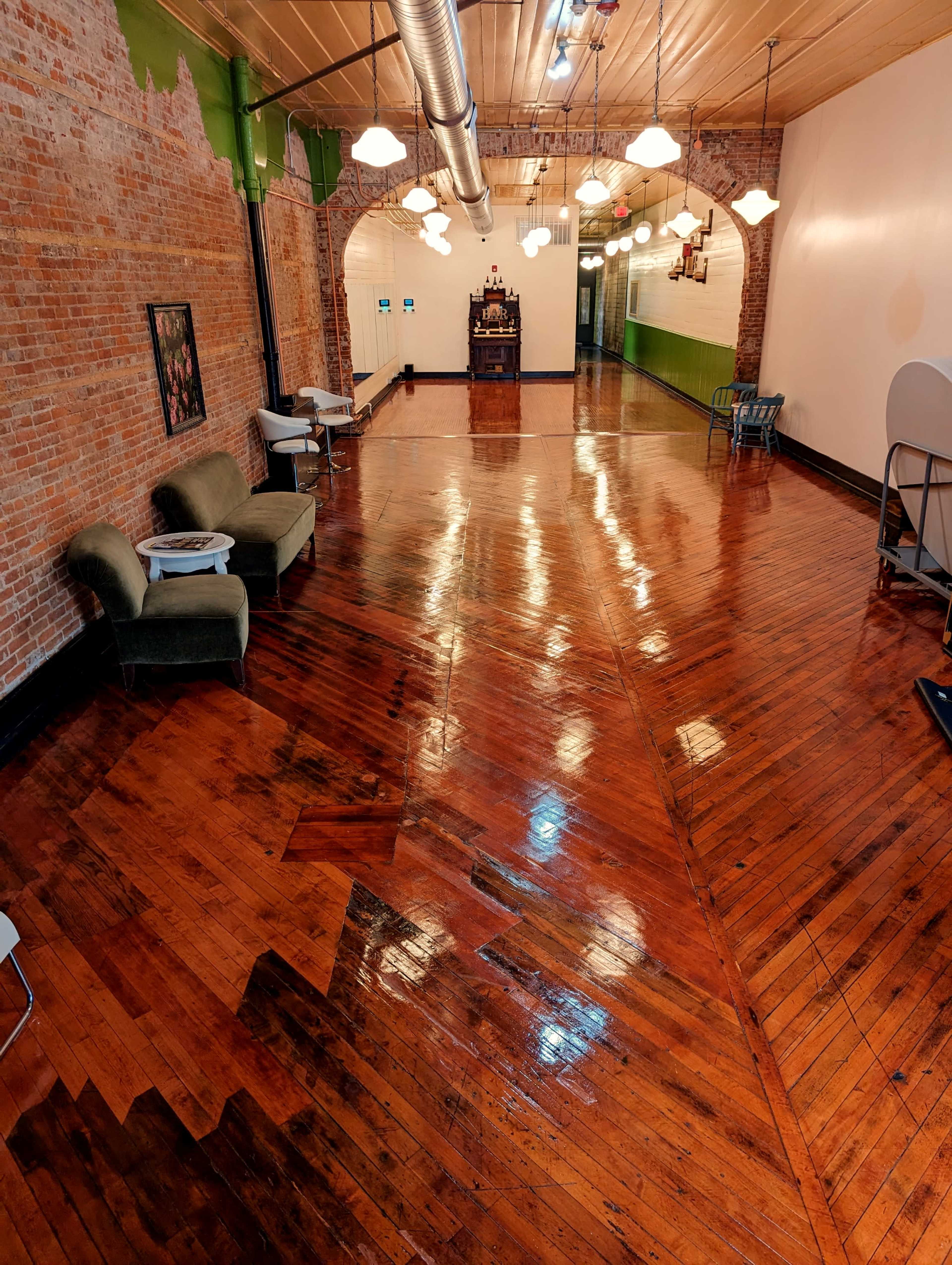 A spacious indoor area with polished wooden floors, exposed brick walls, and green-painted accents, furnished with seating and a bar in the background.