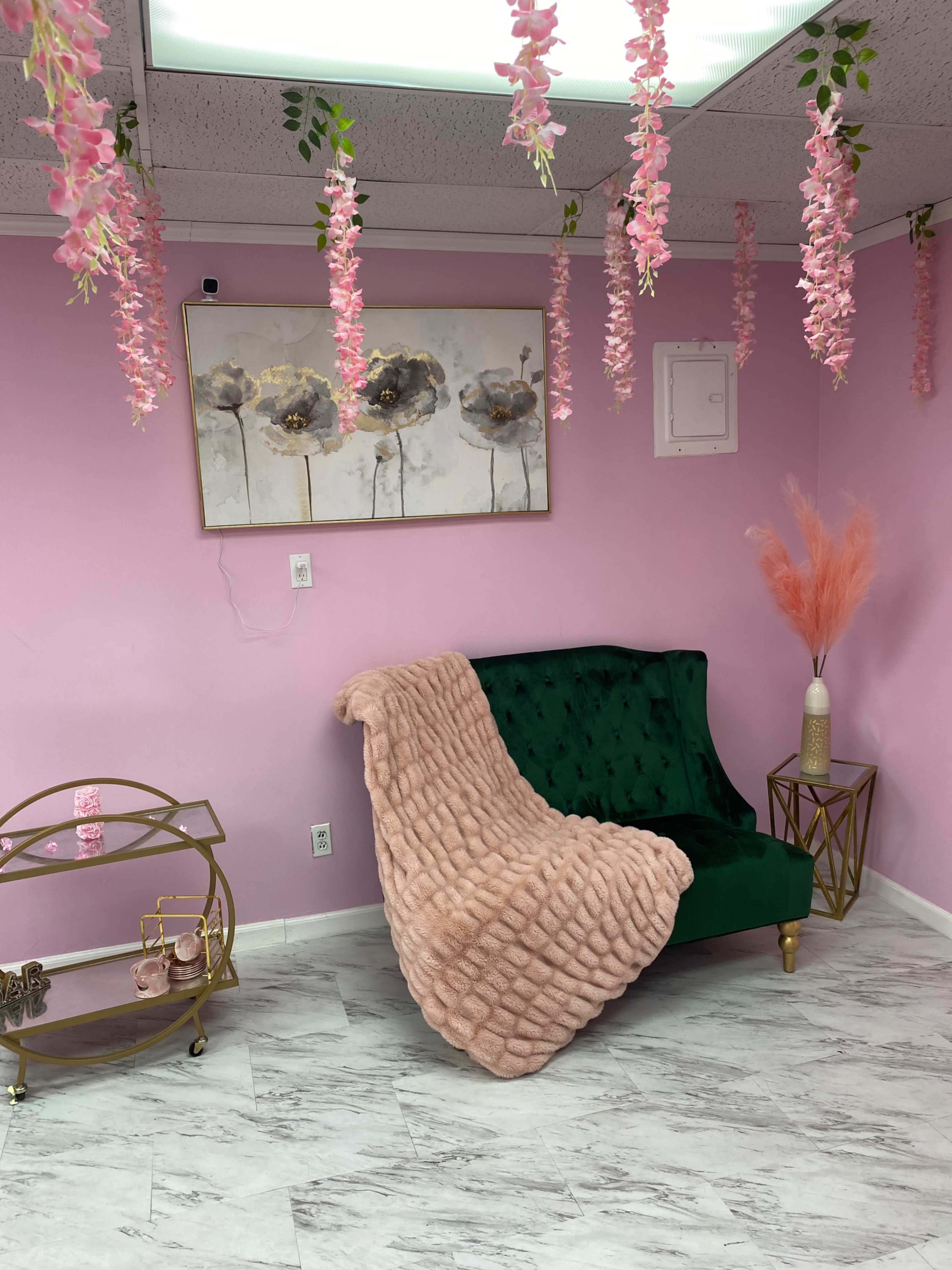 A cozy room with a green velvet loveseat draped with a pink blanket, surrounded by pink floral decorations hanging from the ceiling and a small gold cart in a corner.