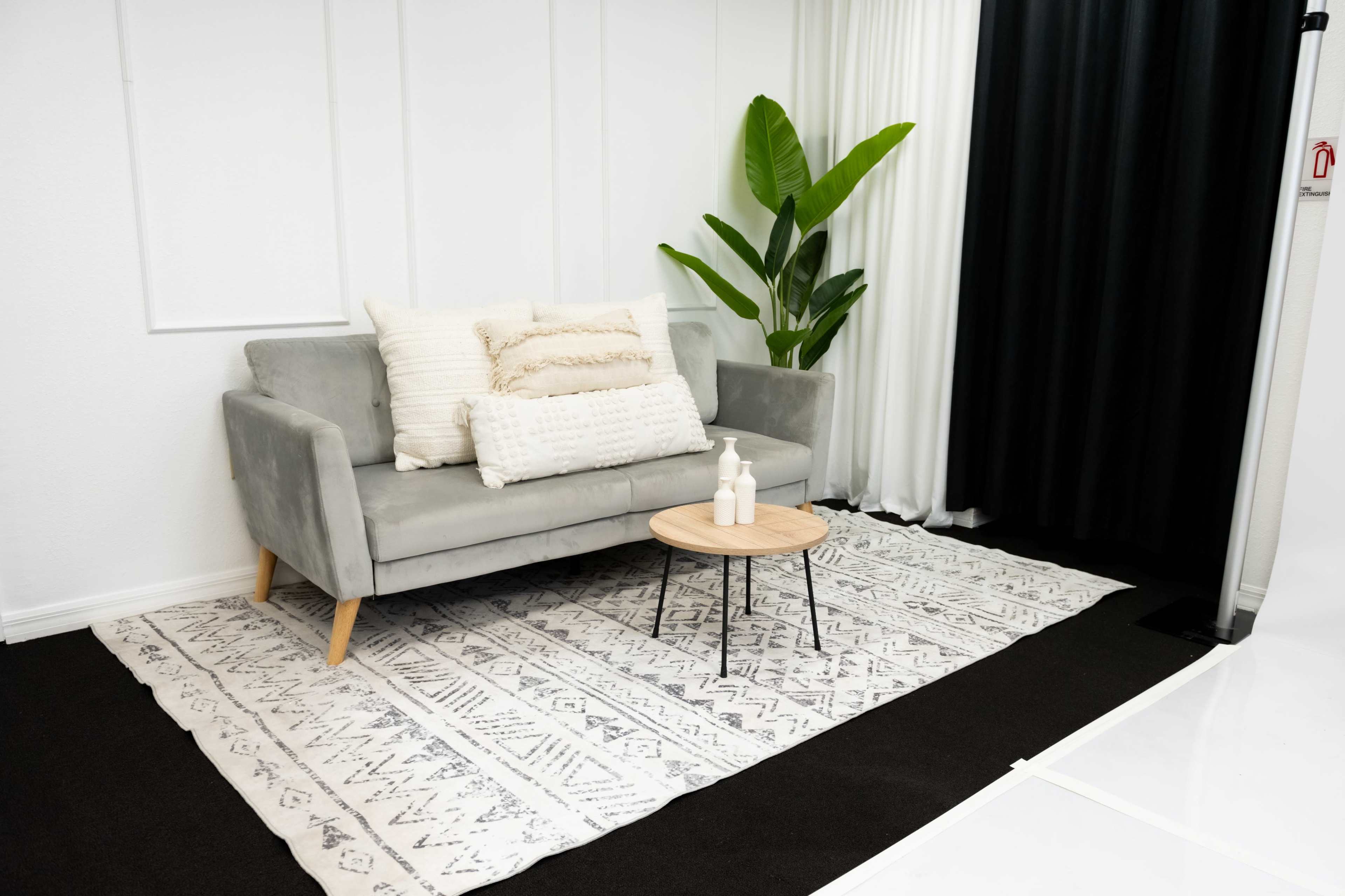 The image shows a modern living space featuring a gray sofa with decorative pillows, a round wooden coffee table, and a large potted plant, set against a white wall and a black curtain.