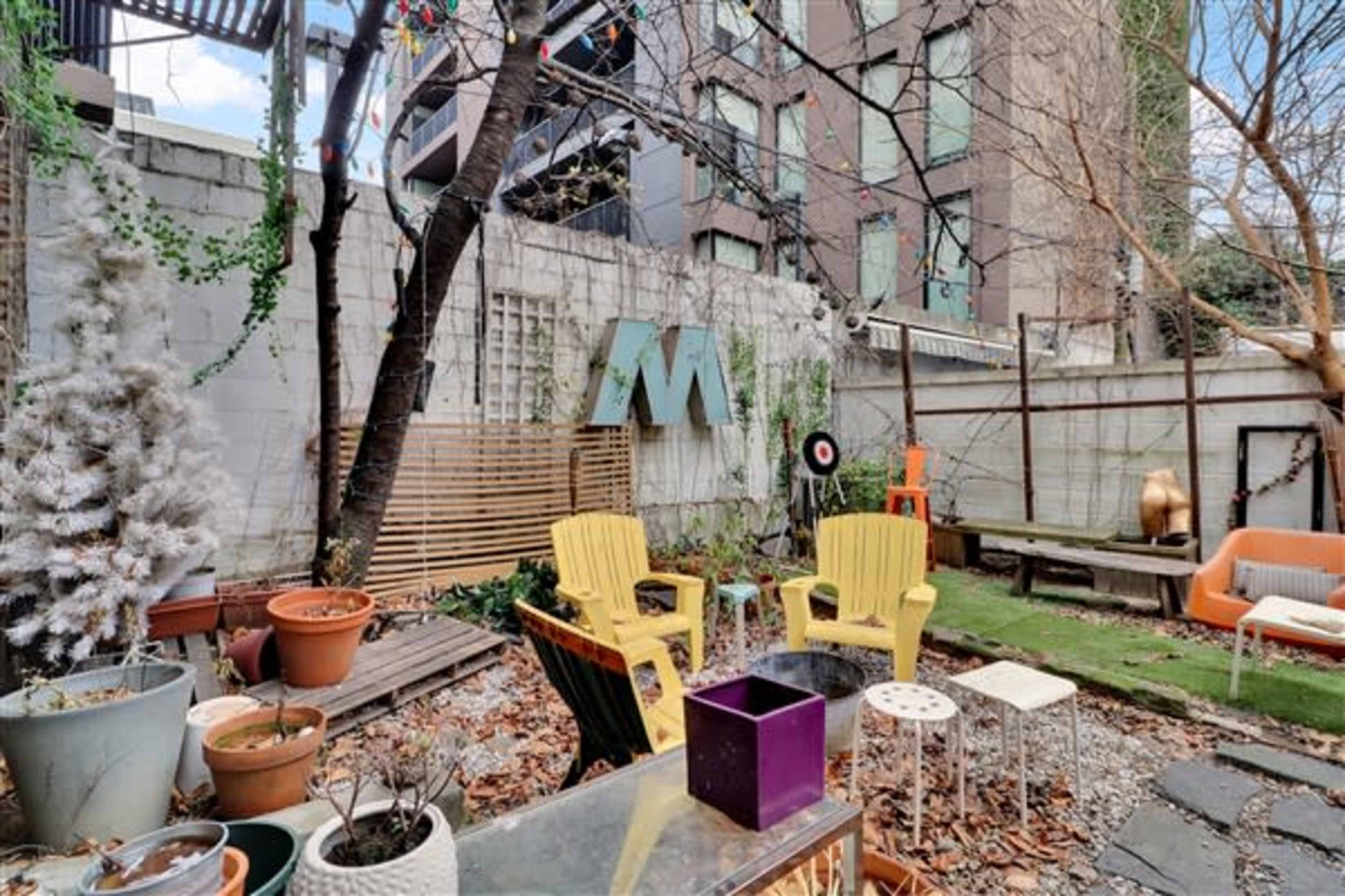 A small garden area filled with various potted plants, colorful chairs, and decorative elements set against a backdrop of modern buildings.