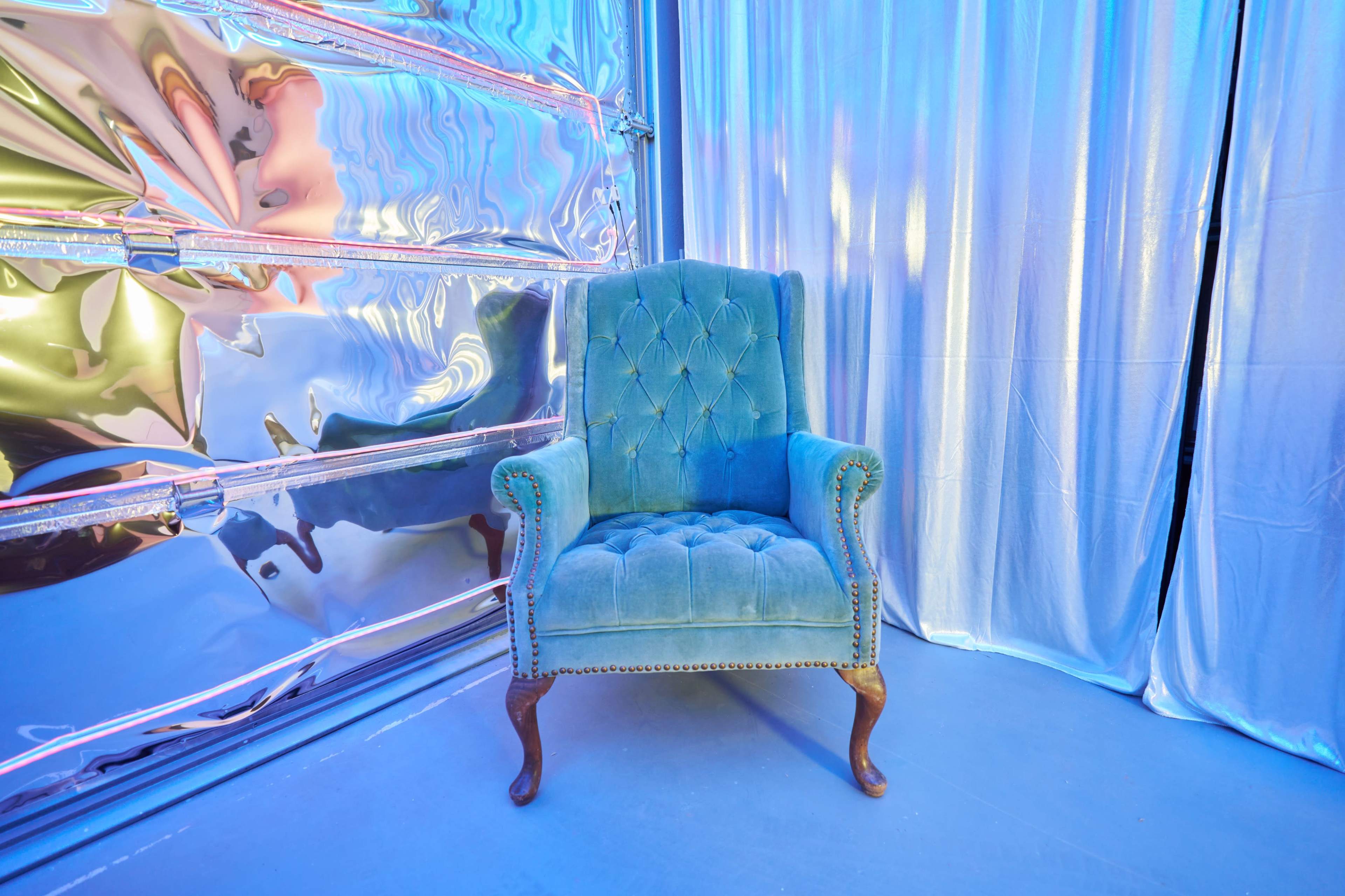 A tufted blue armchair sits against a shiny metallic wall and a silver curtain backdrop.