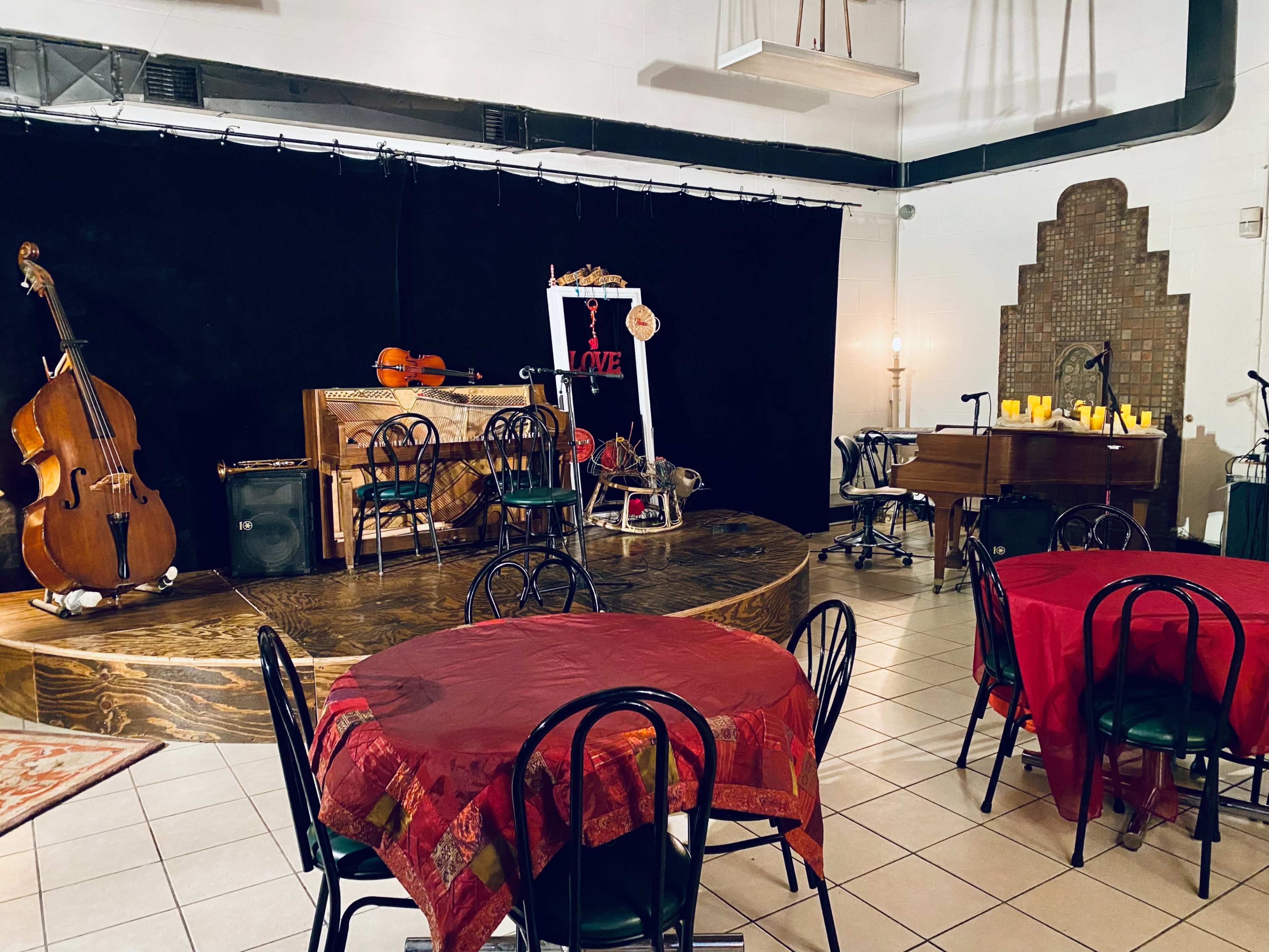 A small stage with musical instruments and a decorated backdrop, surrounded by round tables with red tablecloths in a dimly lit venue.