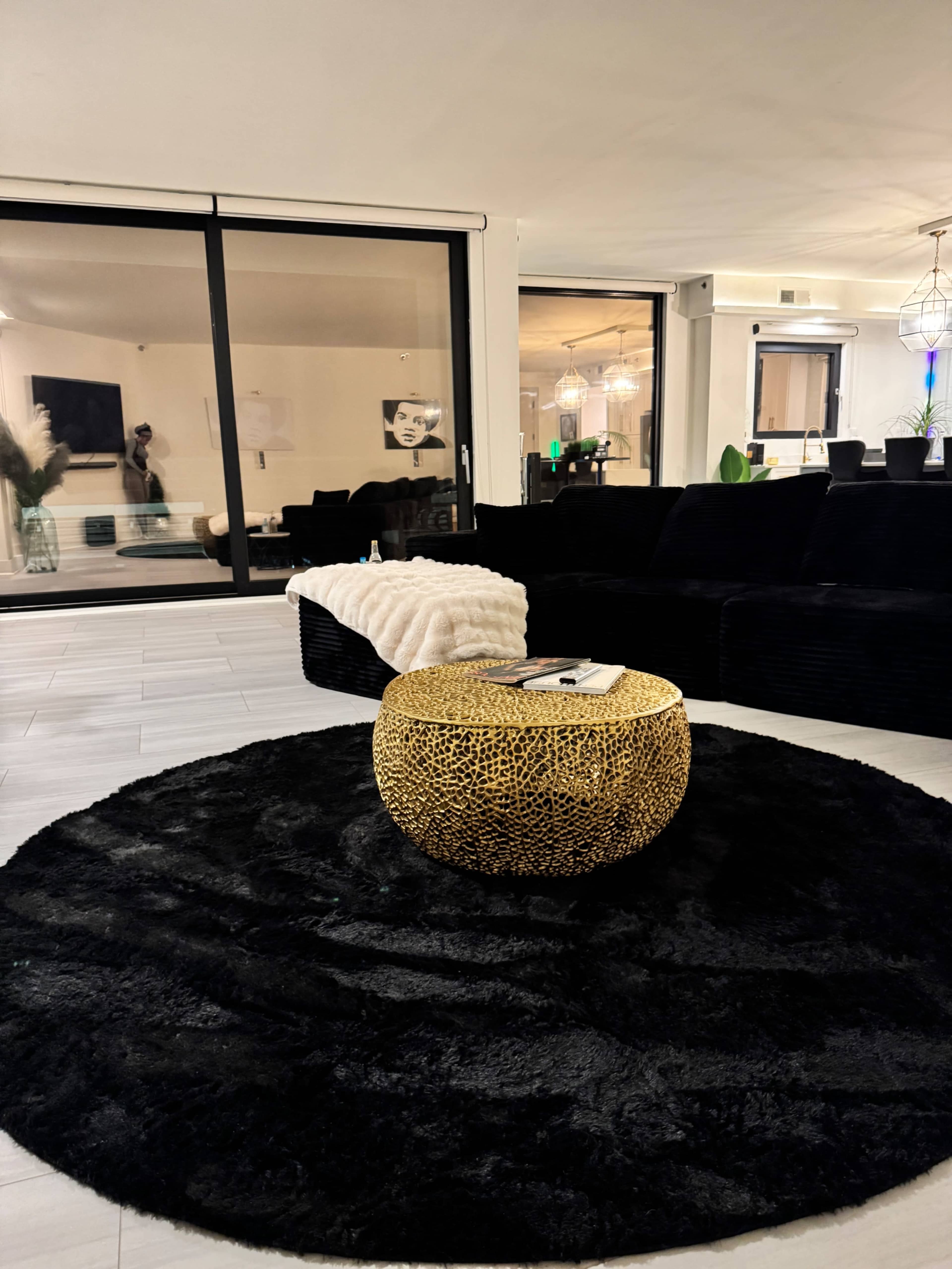 The scene features a modern living room with a black rug, a gold decorative table, and large windows allowing light to enter.