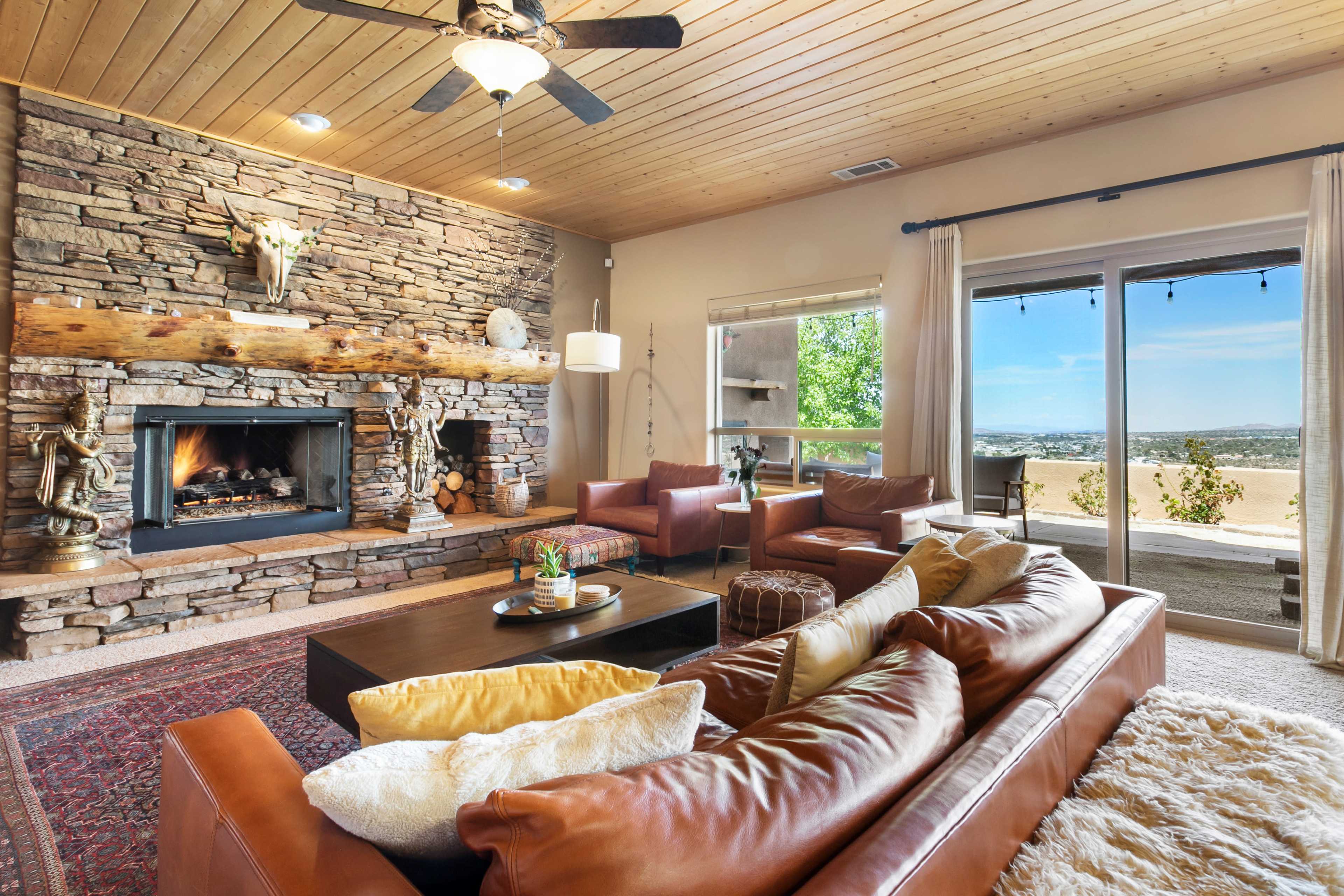 The room features a stone fireplace with a wooden mantel, two red armchairs, a brown leather sofa, and a large window that offers a view of the landscape outside.