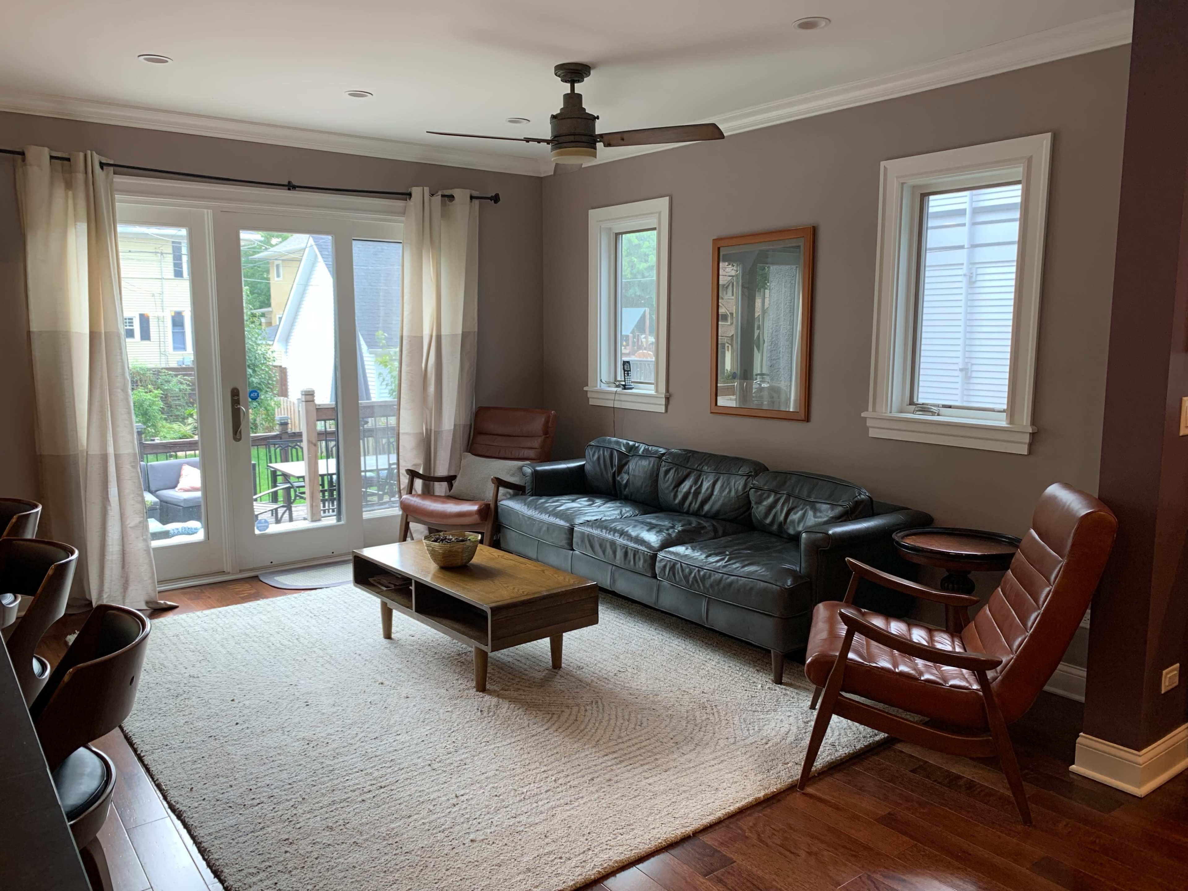 A living room with a dark leather sofa, two brown wooden armchairs, a coffee table, and a view of a deck through sliding glass doors.