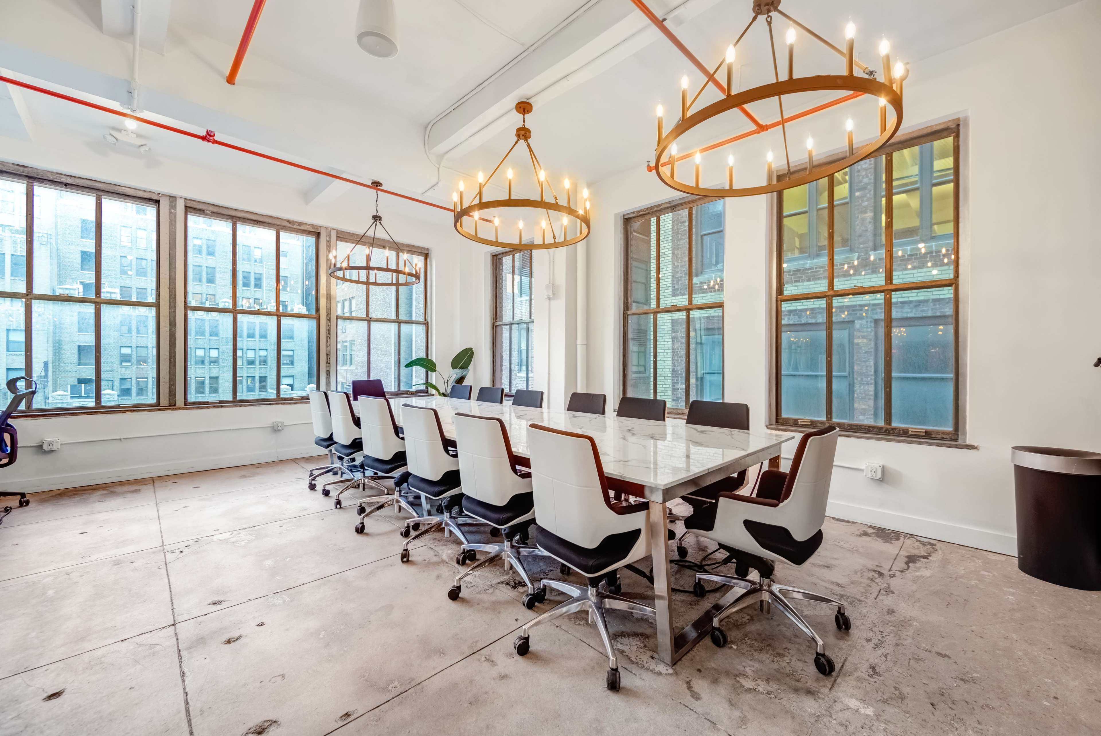A large conference room features a long table surrounded by sleek chairs, illuminated by modern chandeliers and large windows.