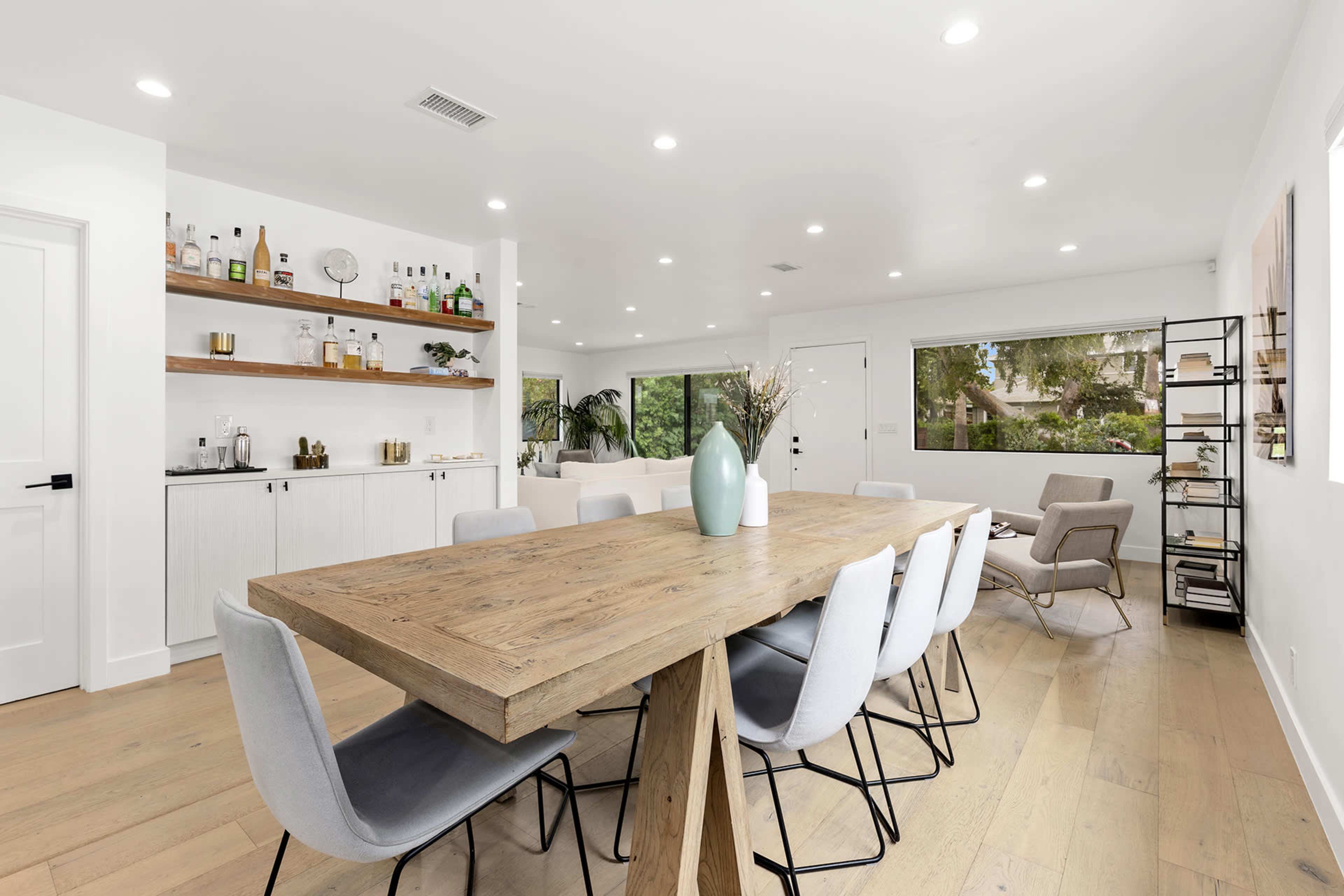 A spacious dining area features a large wooden table surrounded by light-colored chairs, with a modern kitchen and living space visible in the background.