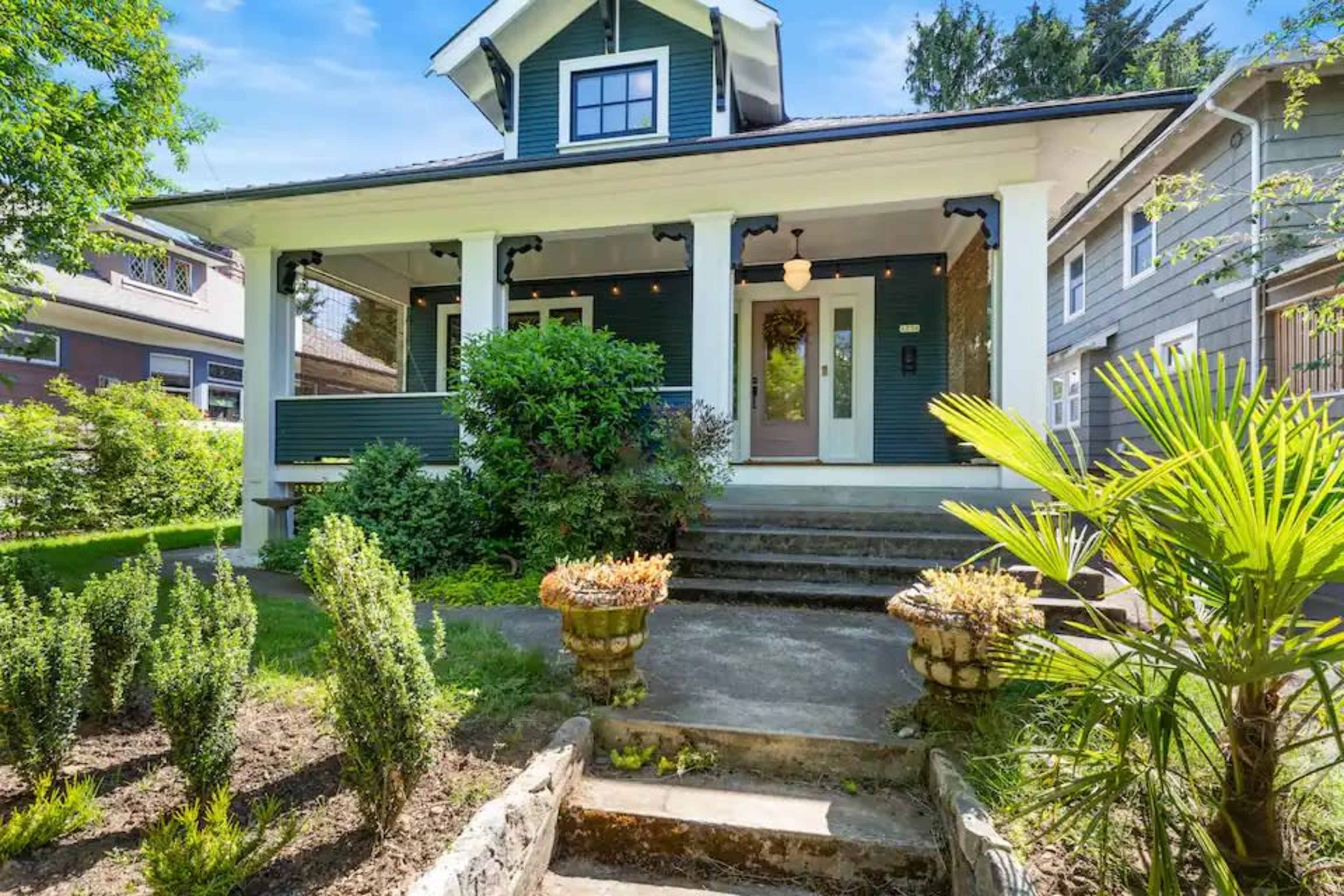 A charming green house with a front porch and a well-maintained garden leading up to the entrance.