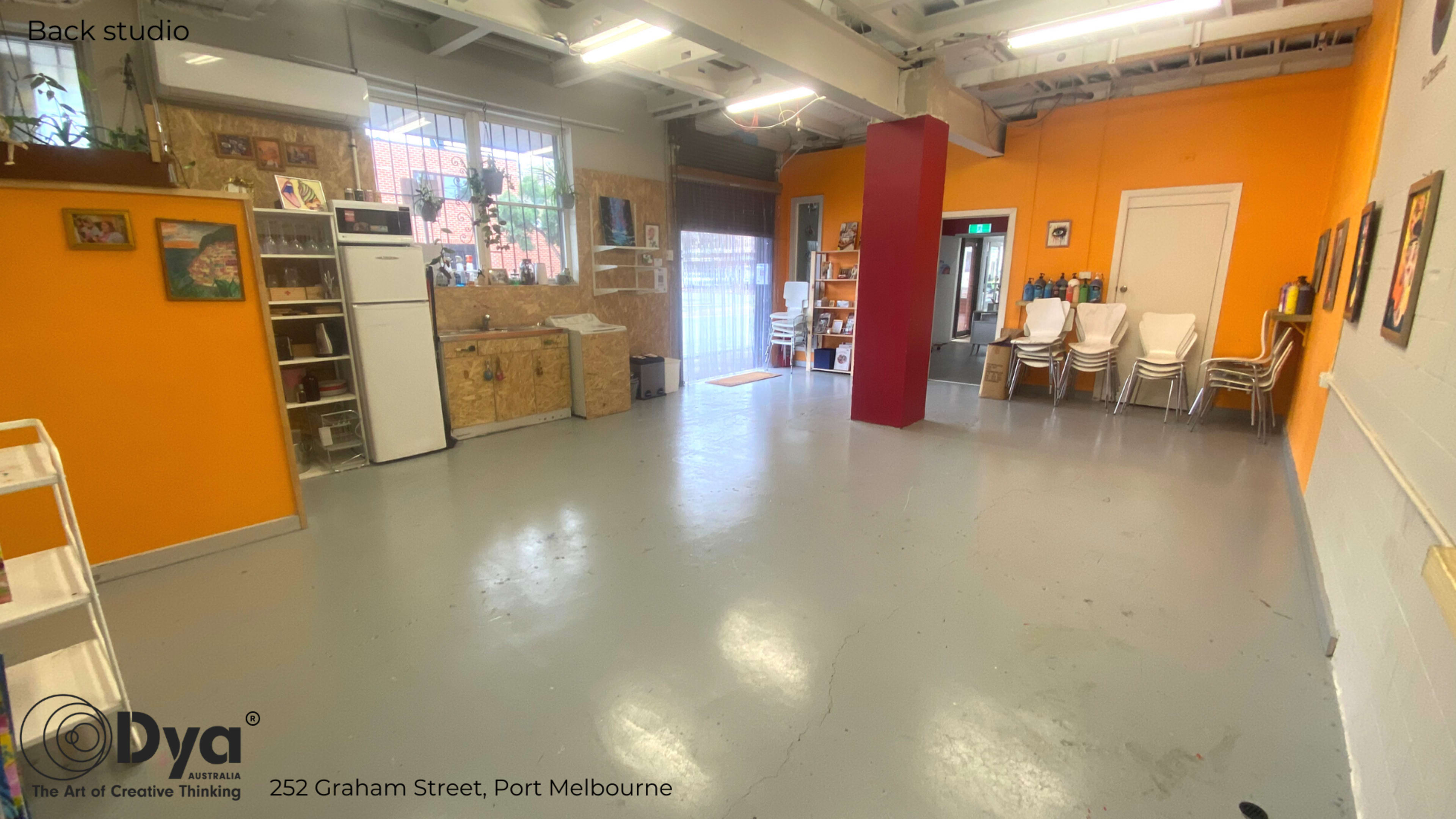 An empty studio space with orange and white walls, a large window, and various furniture items including a refrigerator and stacked chairs.