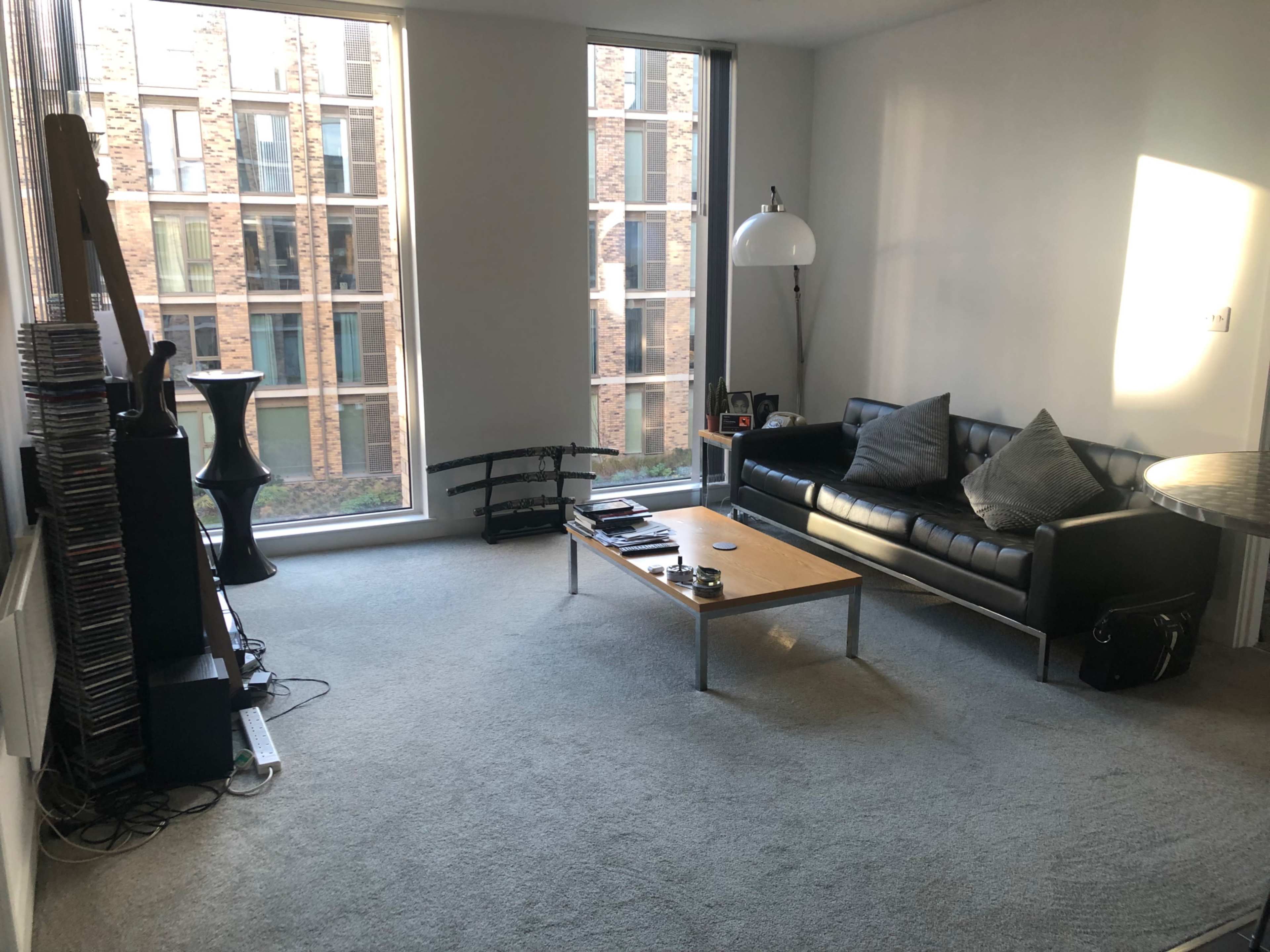 The image shows a sparsely decorated living room with a black leather sofa, a wooden coffee table, and two large windows revealing a view of a brick building outside.
