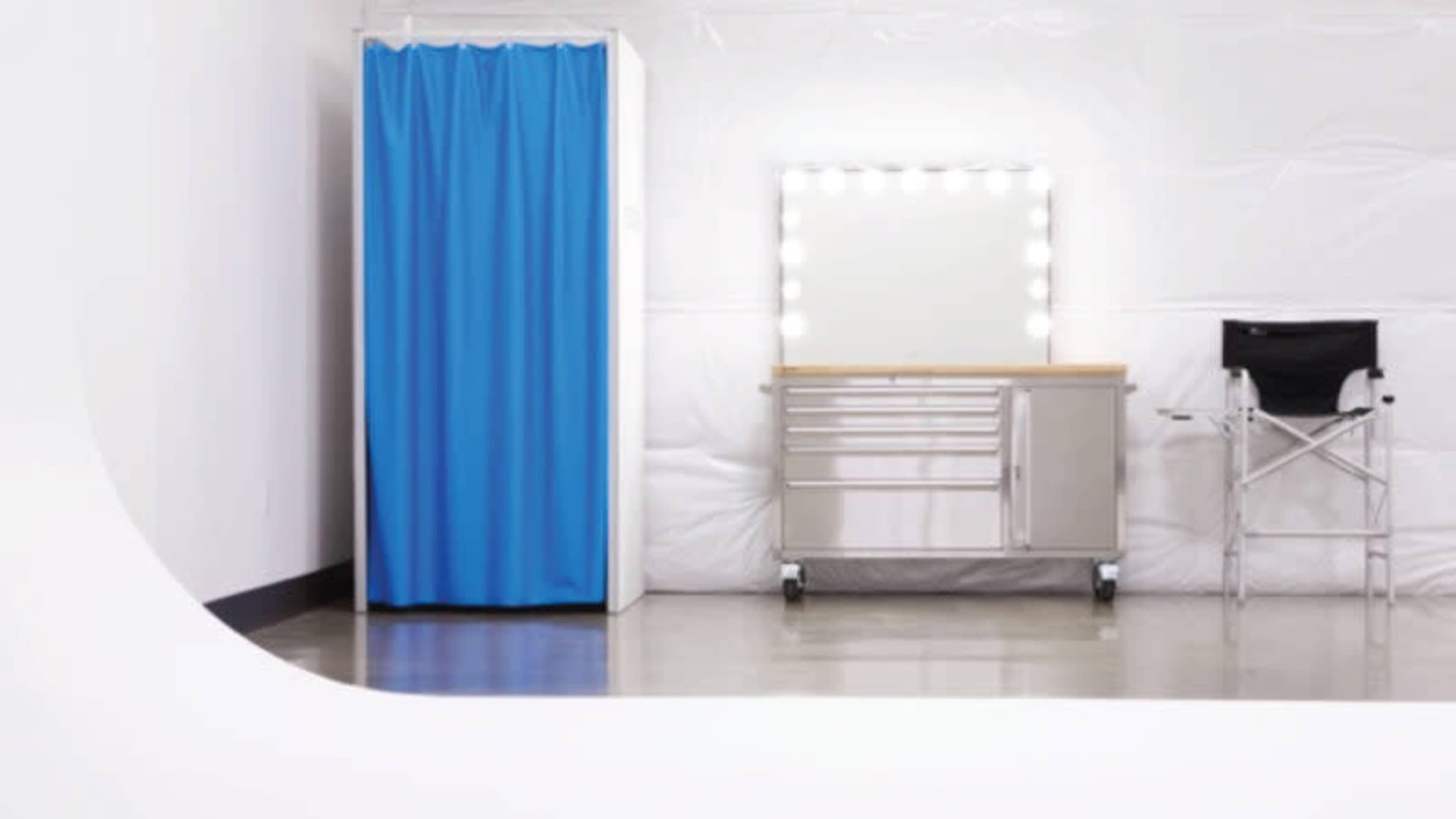 A room with a blue curtain for privacy, a bright mirror with lights, and a silver cart next to a black director's chair.