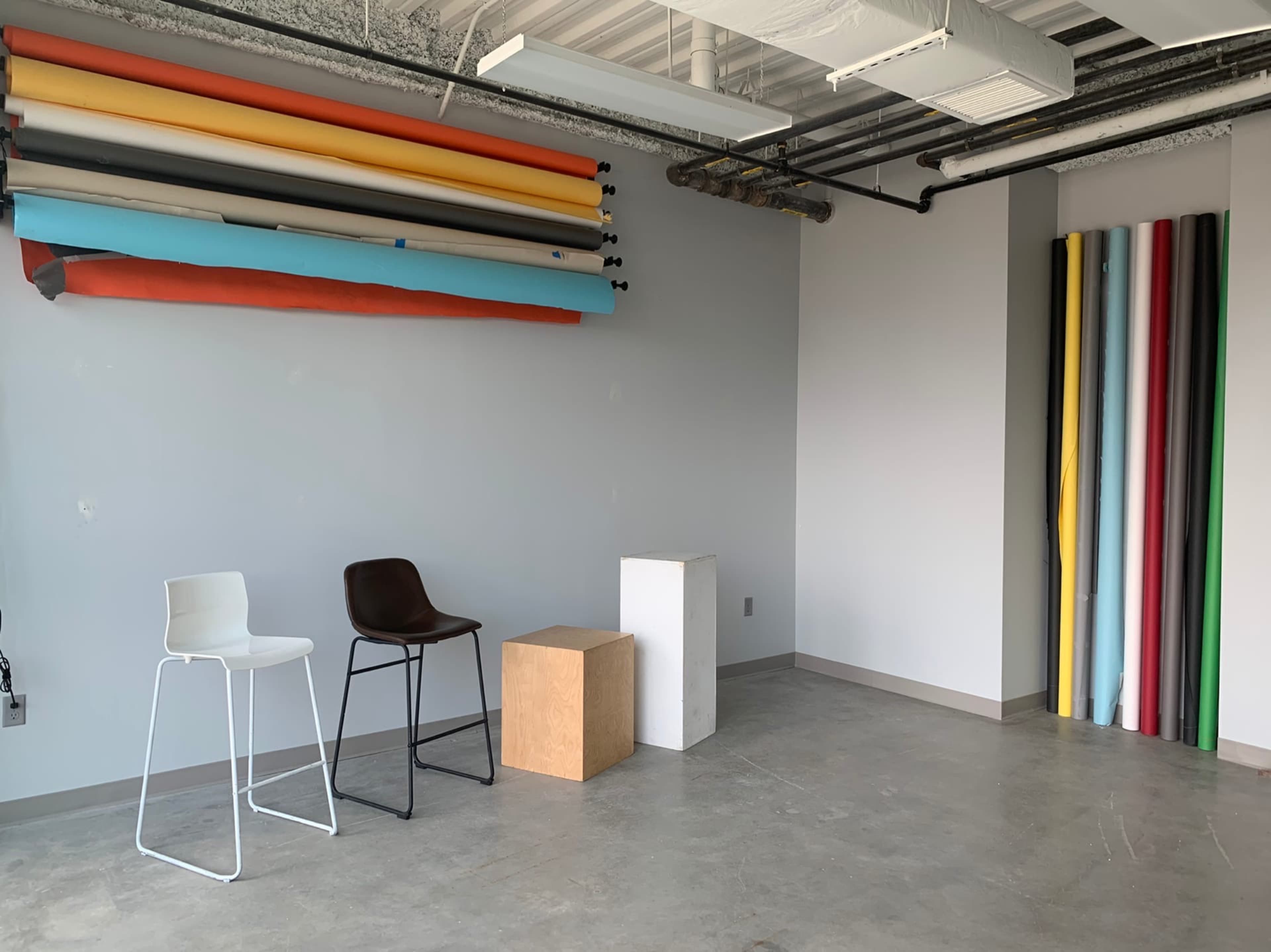 A minimalist studio space with colored backdrop rolls mounted on the wall and three different types of stools and a cube placed on the floor.