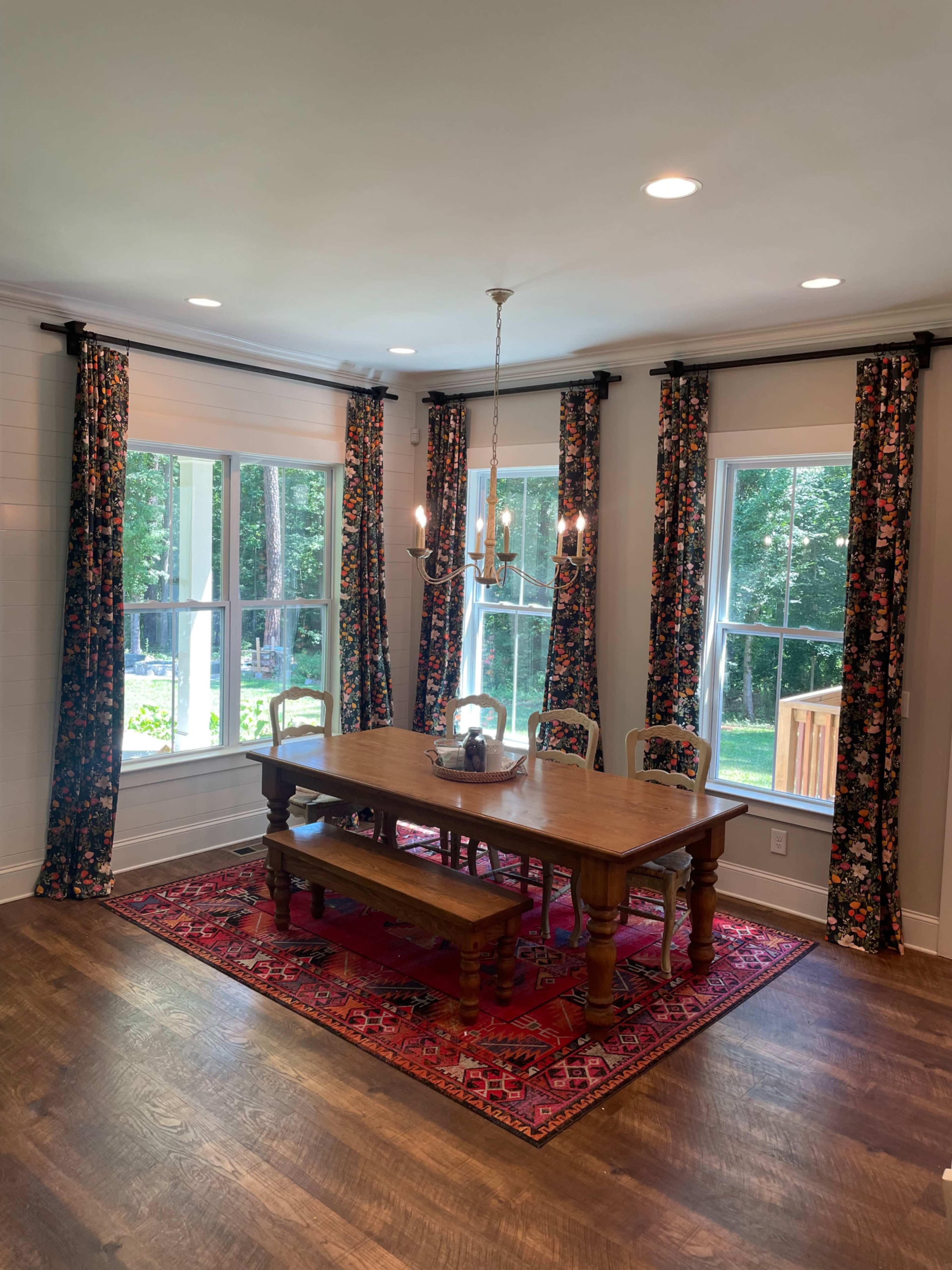 A dining room features a wooden table surrounded by chairs, with floral curtains framing large windows that overlook a green backyard.