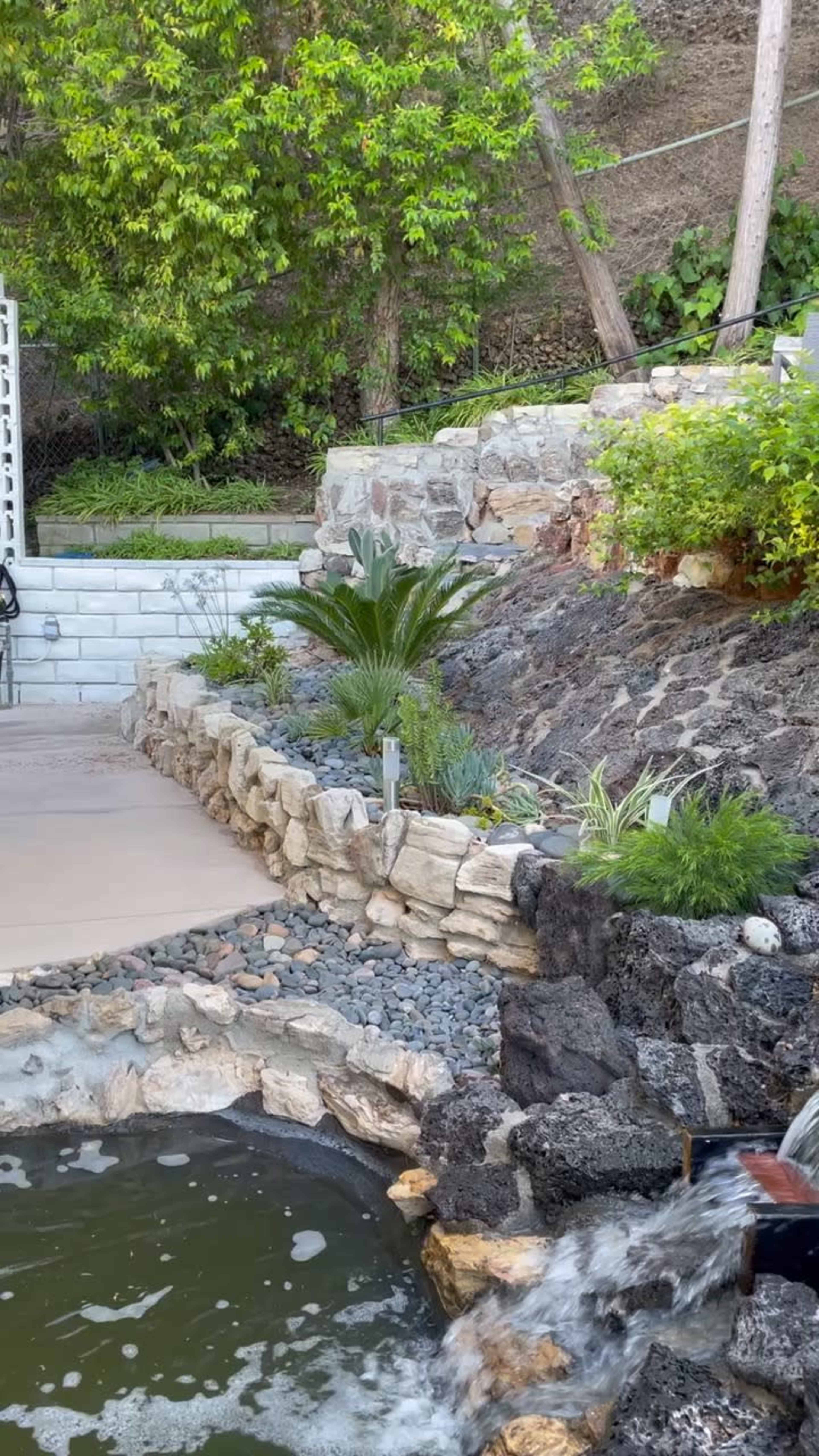 A waterfall flows into a small pond surrounded by stone landscaping and various plants.