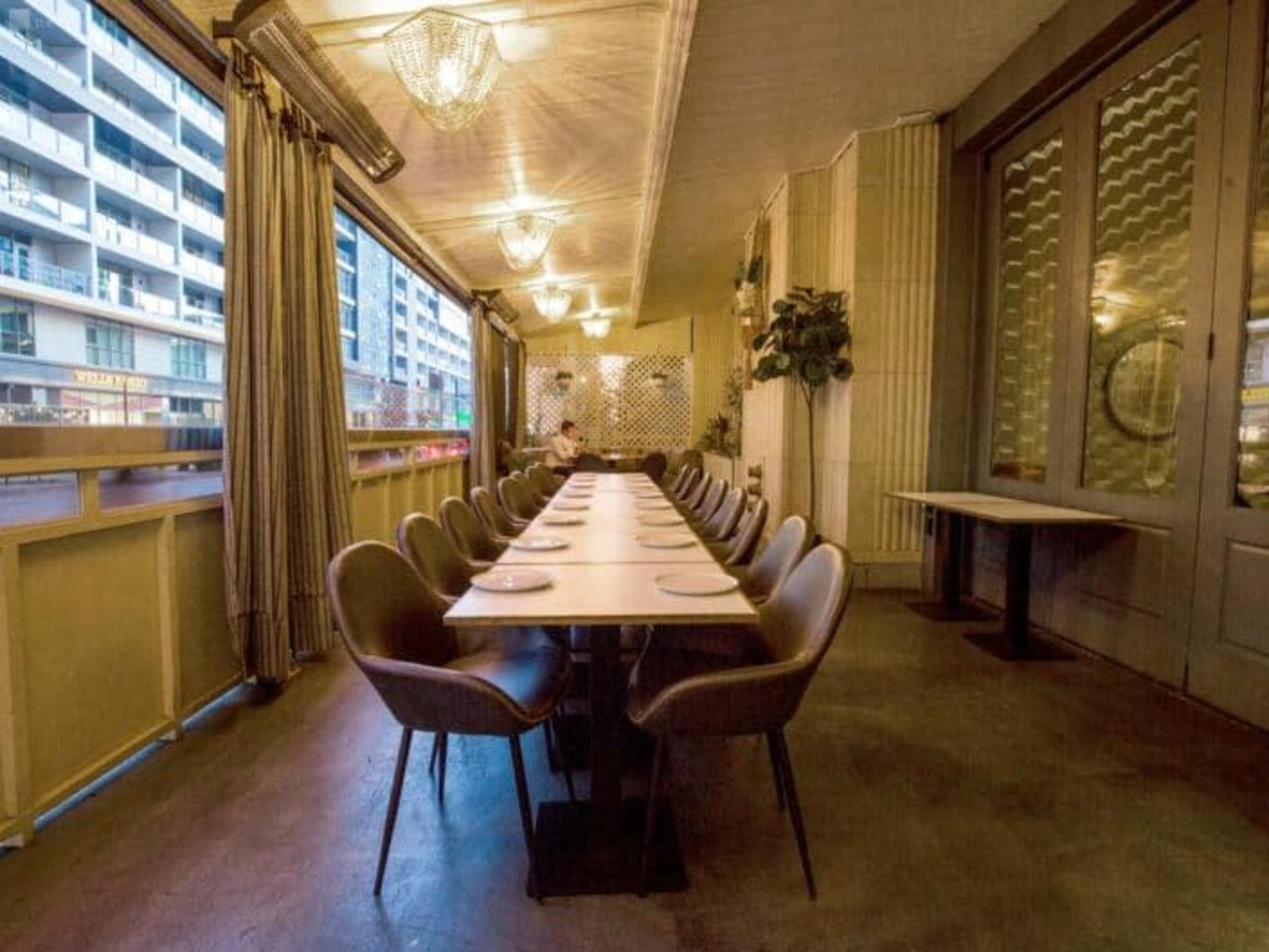 A long, empty dining table with chairs is set in a well-lit restaurant space that features large windows and modern decor.