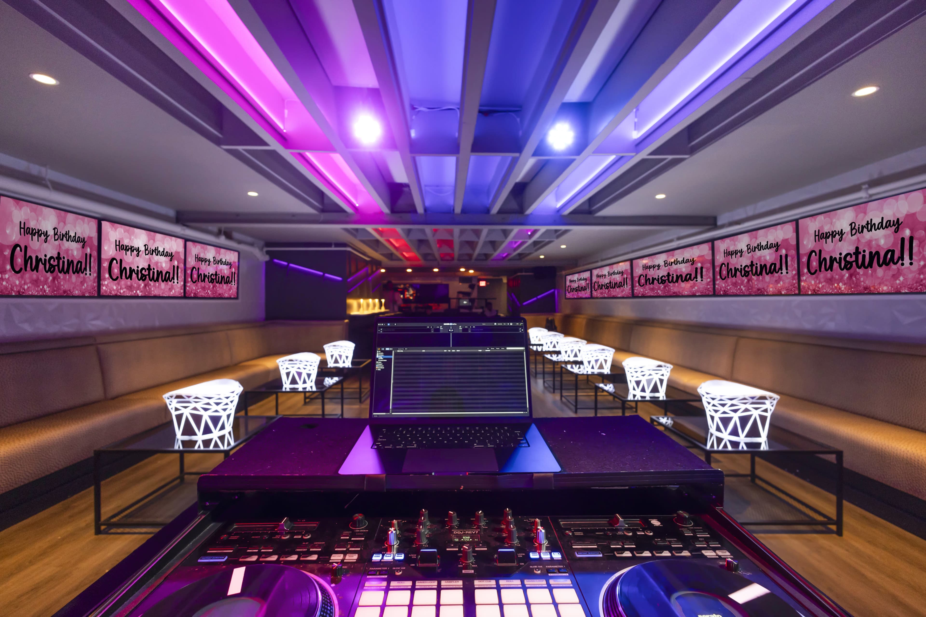 A DJ booth is set up in a brightly lit venue decorated with "Happy Birthday Christina!" banners and illuminated chairs.