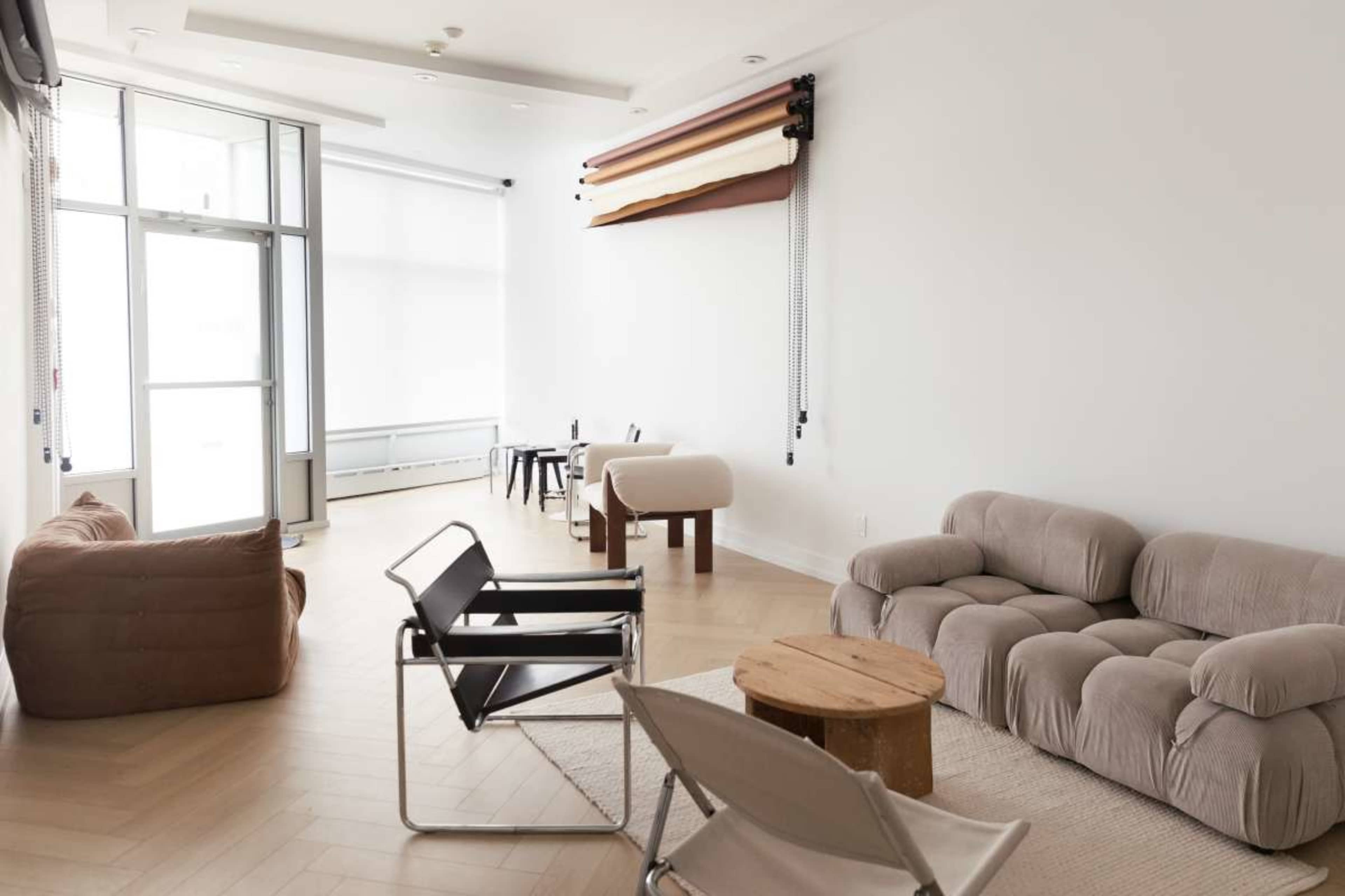 The image shows a modern, minimally furnished interior space with various seating arrangements, including a large couch, armchairs, and small tables.
