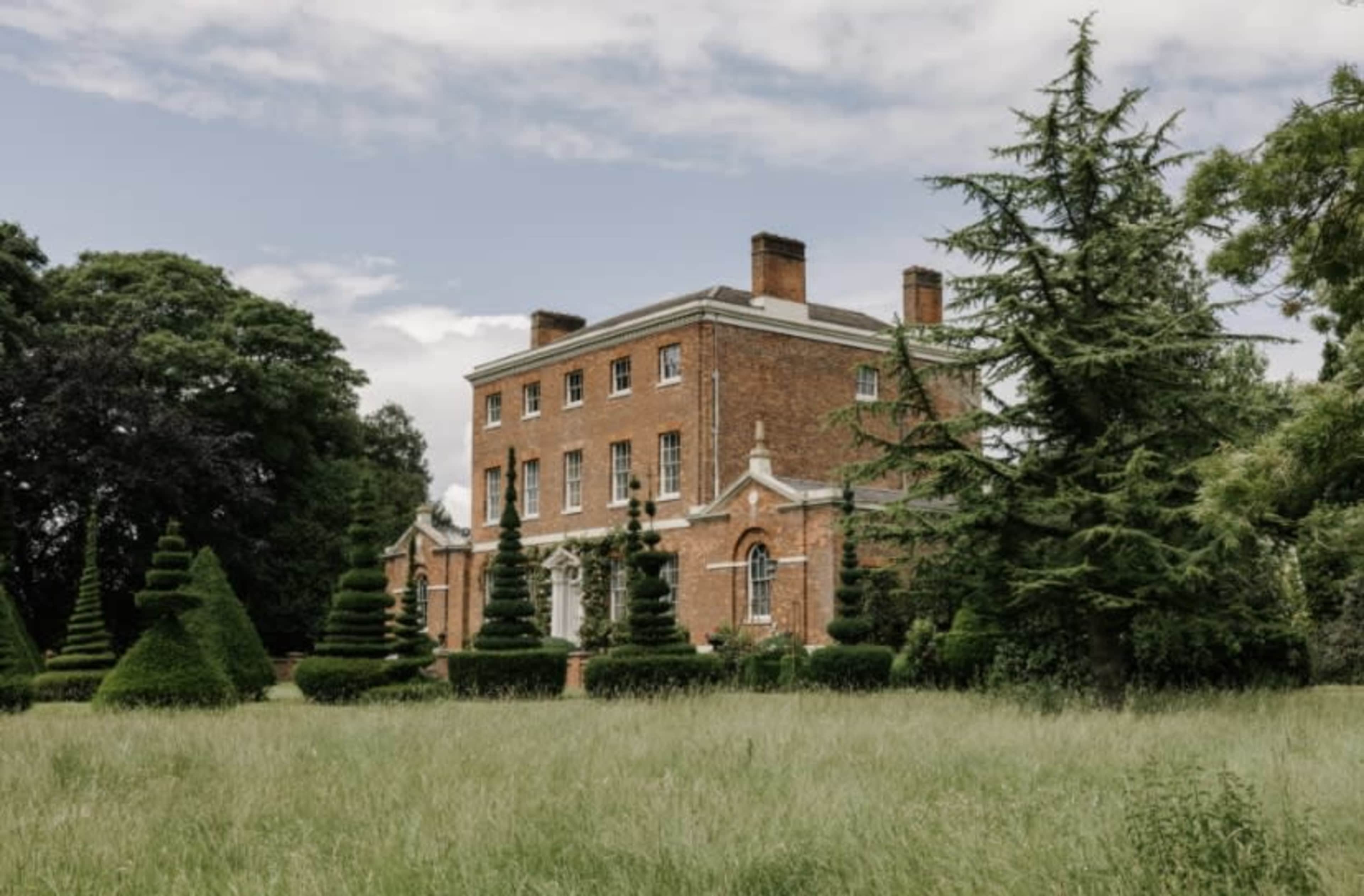 A large, red-brick mansion is surrounded by neatly trimmed topiary and a grassy lawn under a cloudy sky.