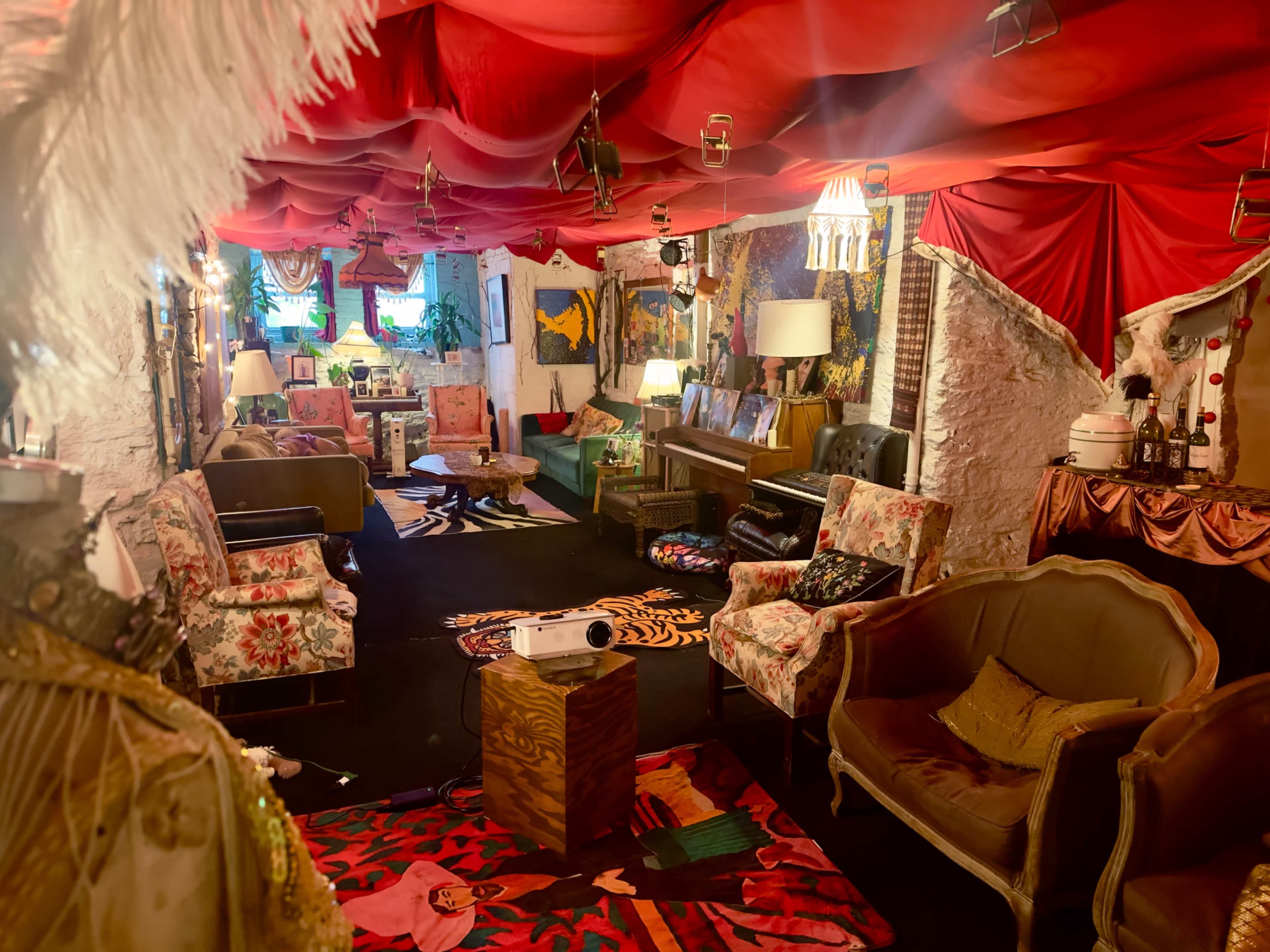 The image shows a vibrant, eclectic living space with colorful draped fabric, vintage furniture, and various art pieces adorning the walls.