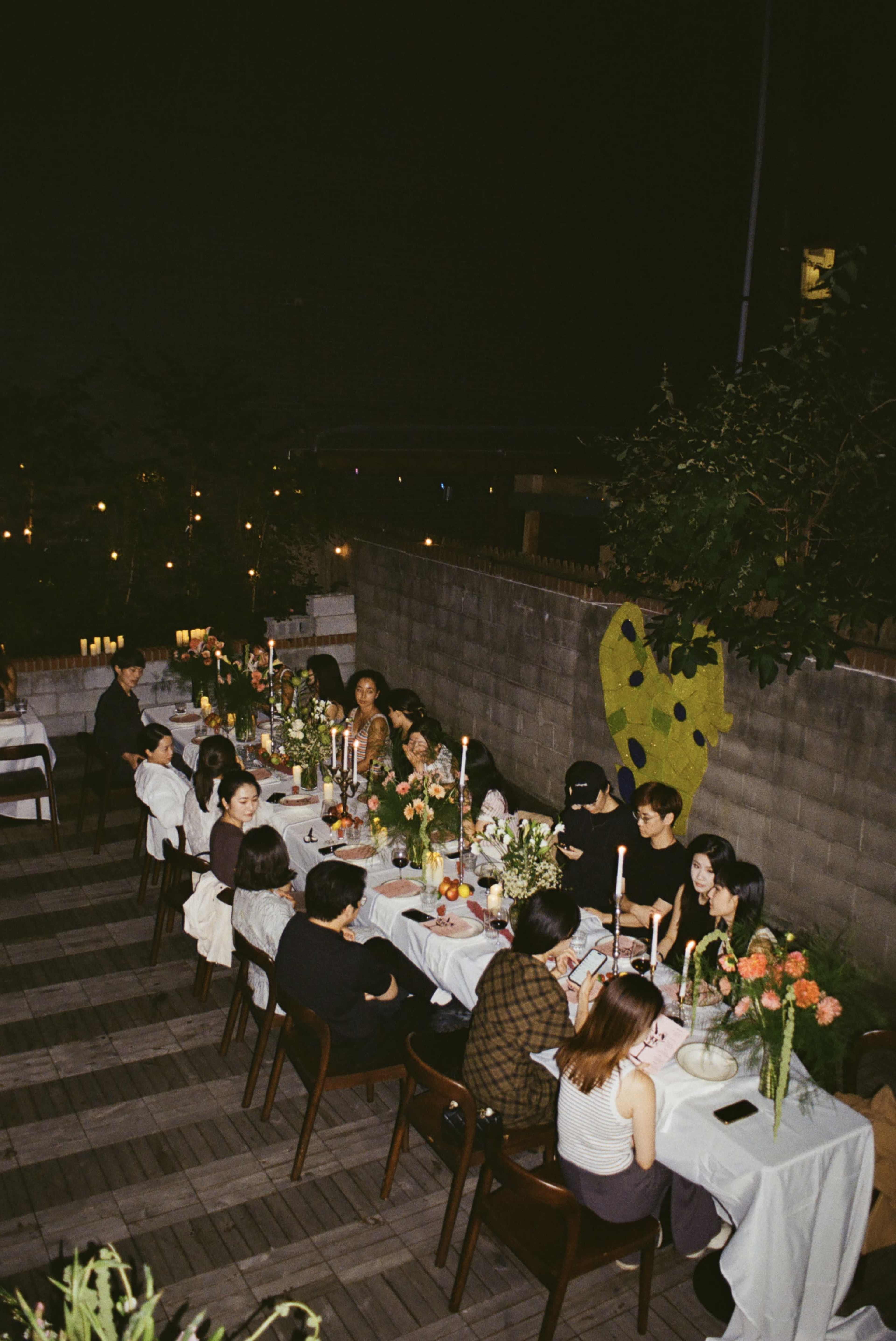 A group of people sits at a long table adorned with flowers and candles, enjoying an evening gathering in a well-lit outdoor space.