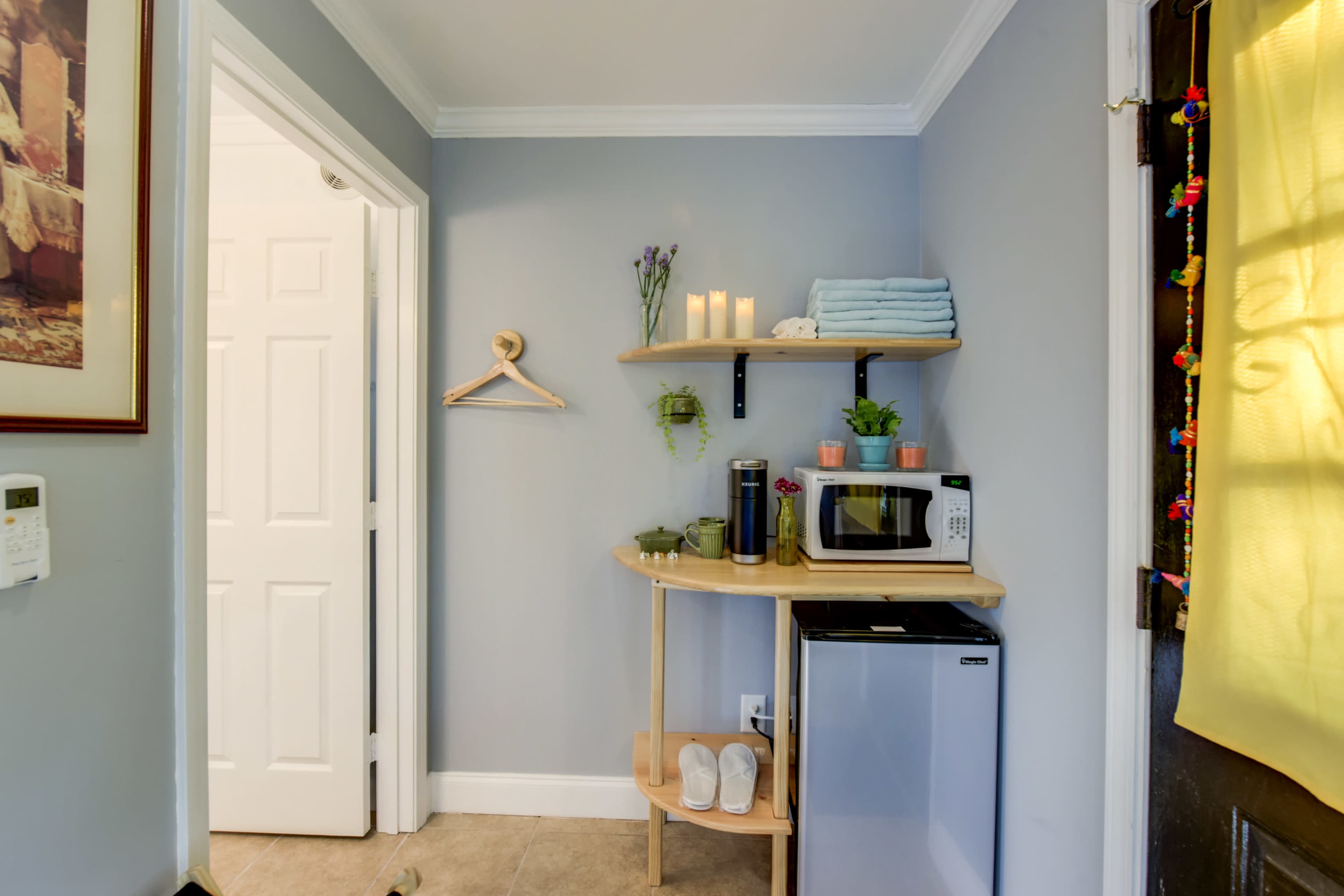 A small kitchen corner features a microwave, a mini fridge, and open shelves with stacked towels and decorative plants, alongside a door leading to another room.