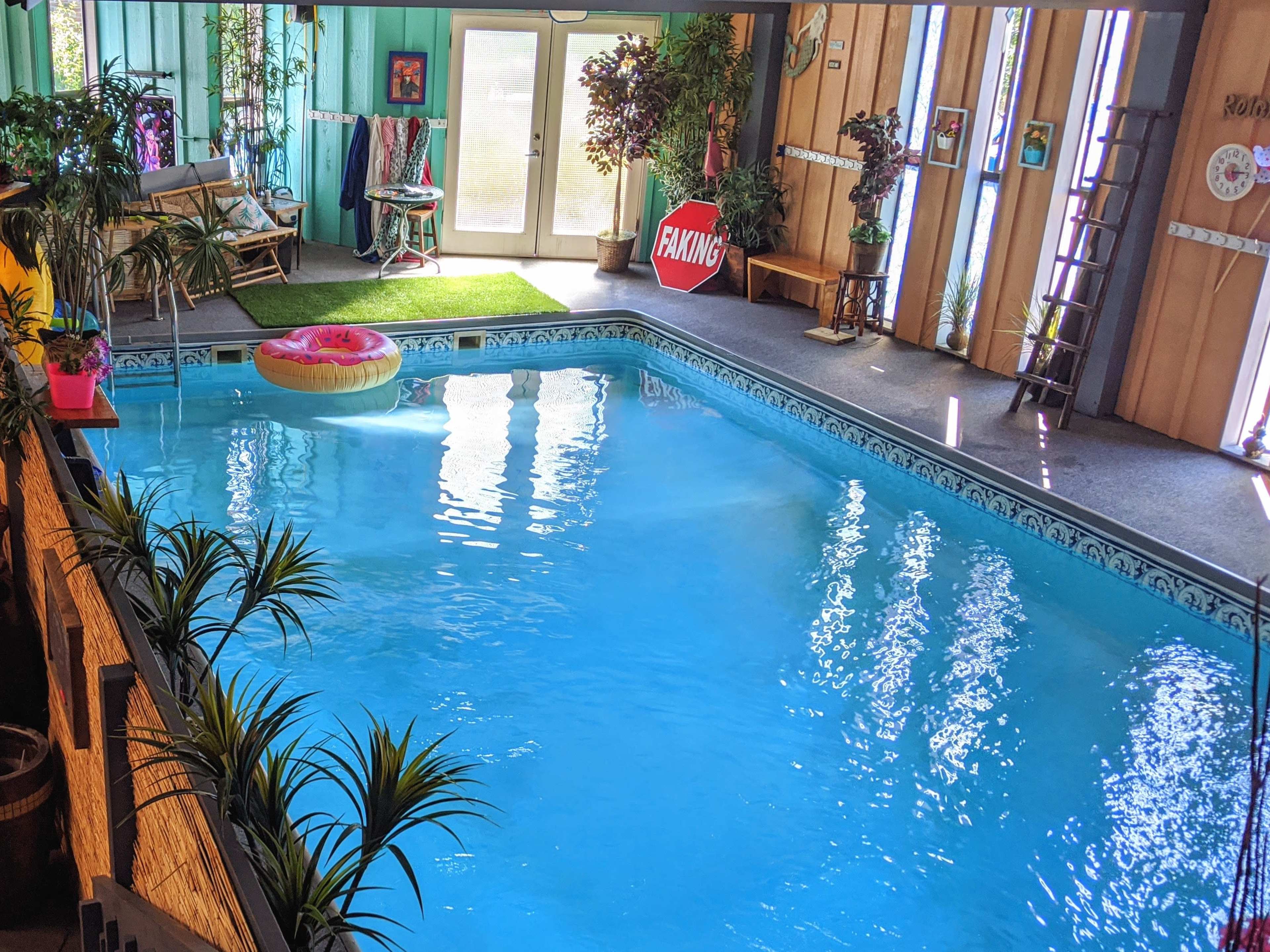 An indoor swimming pool surrounded by plants, a pool float, and lounge chairs, with large windows allowing natural light to enter.