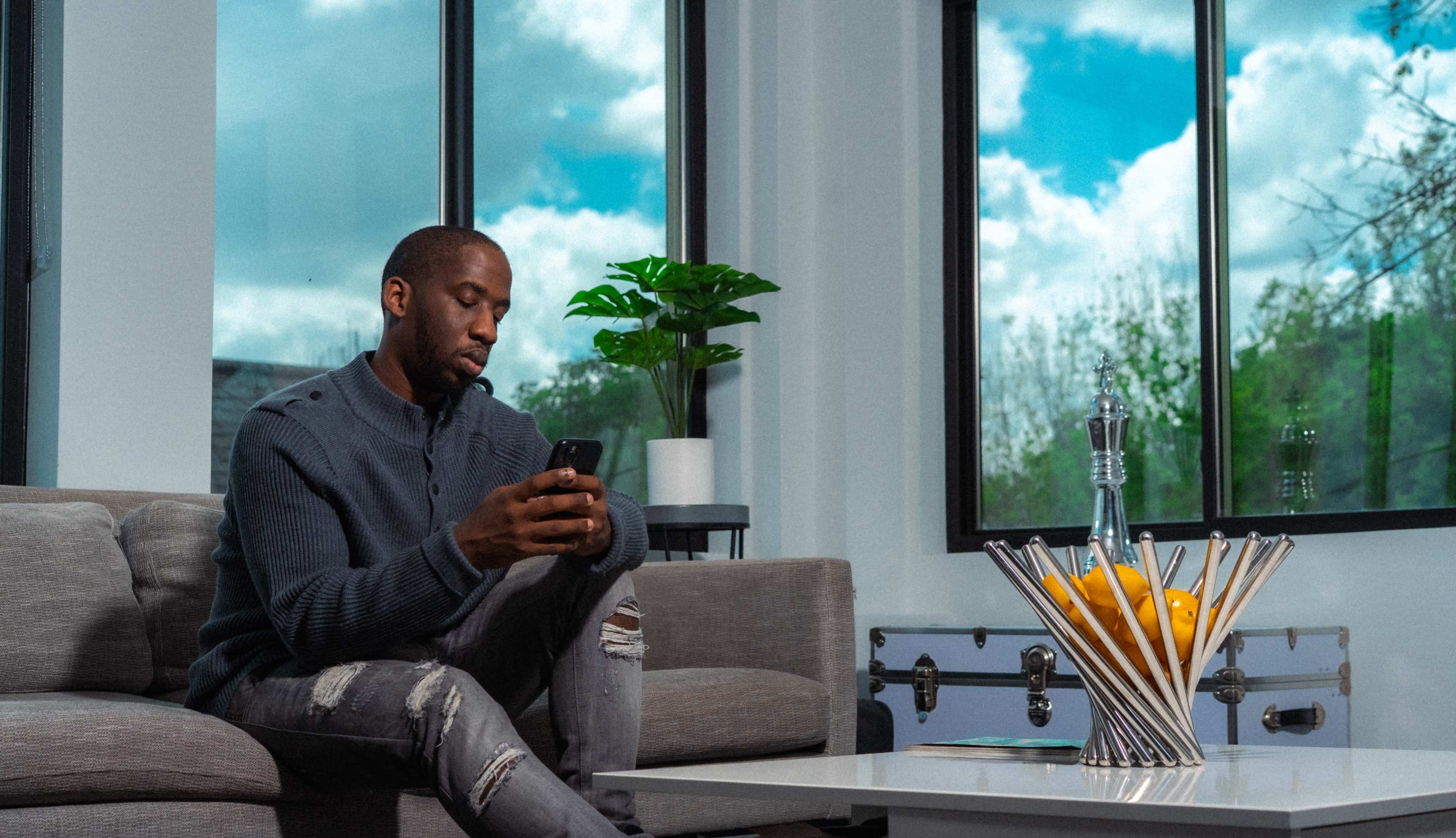 A man sits on a grey couch, looking at his phone, while a plant and a fruit bowl are placed on a table beside him, with large windows showing a blue sky outside.