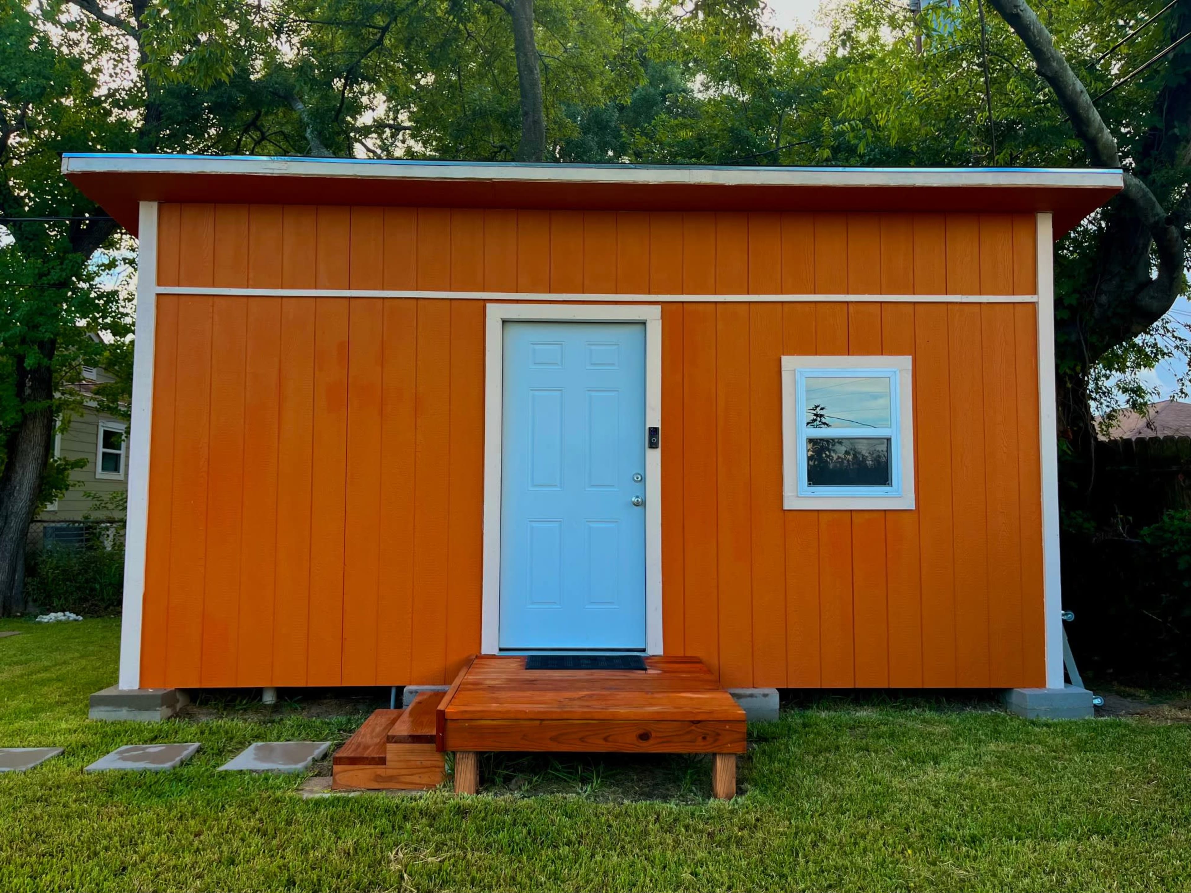A small orange wooden house with a light blue door and a window, set on a grassy area.