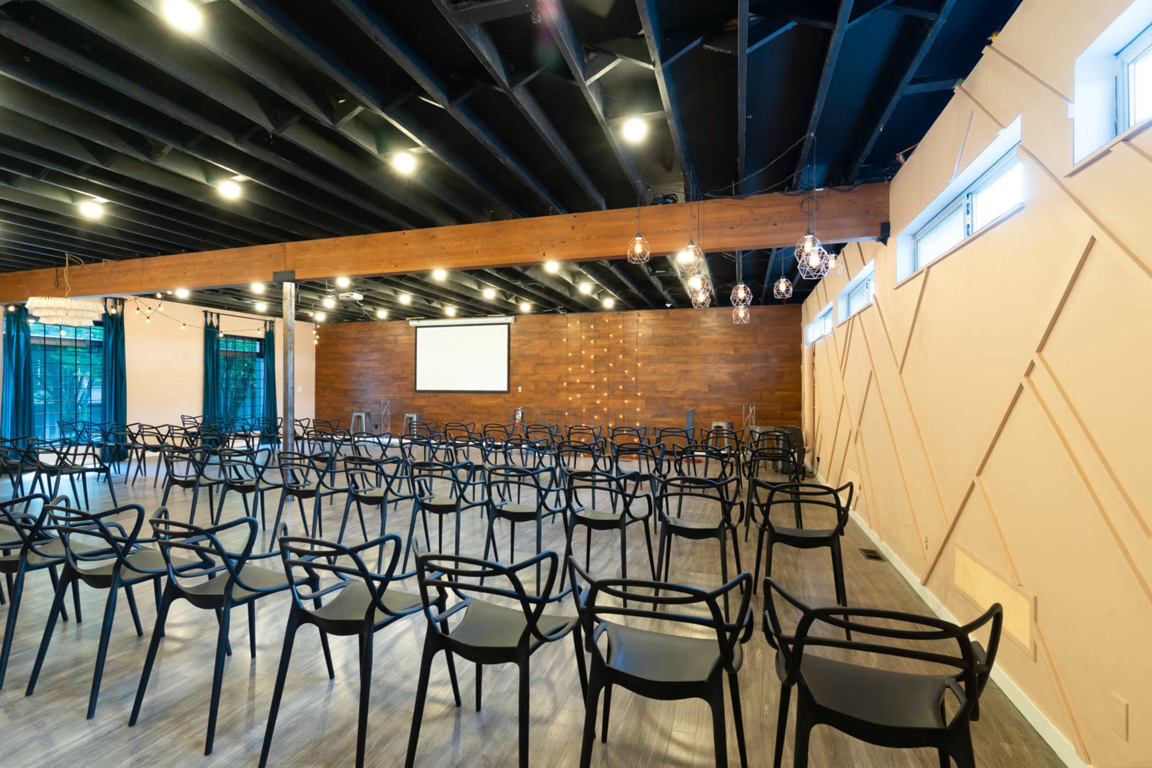 A conference room is set up with rows of black chairs facing a projector screen, surrounded by wooden paneling and modern lighting.
